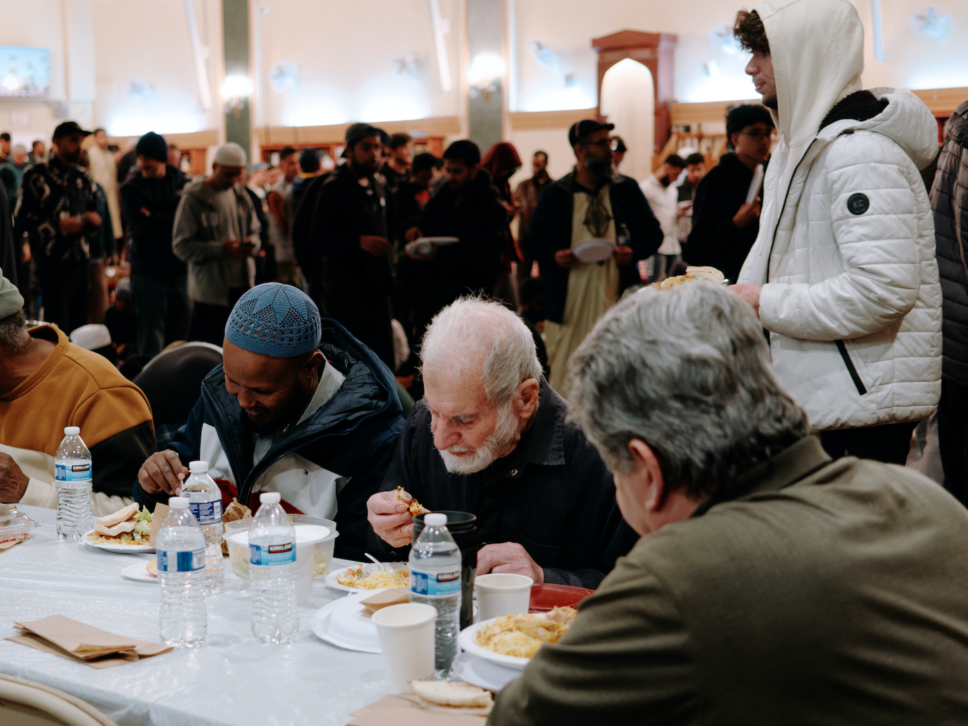 Men sit at a table and eat in front of lines of men waiting for food