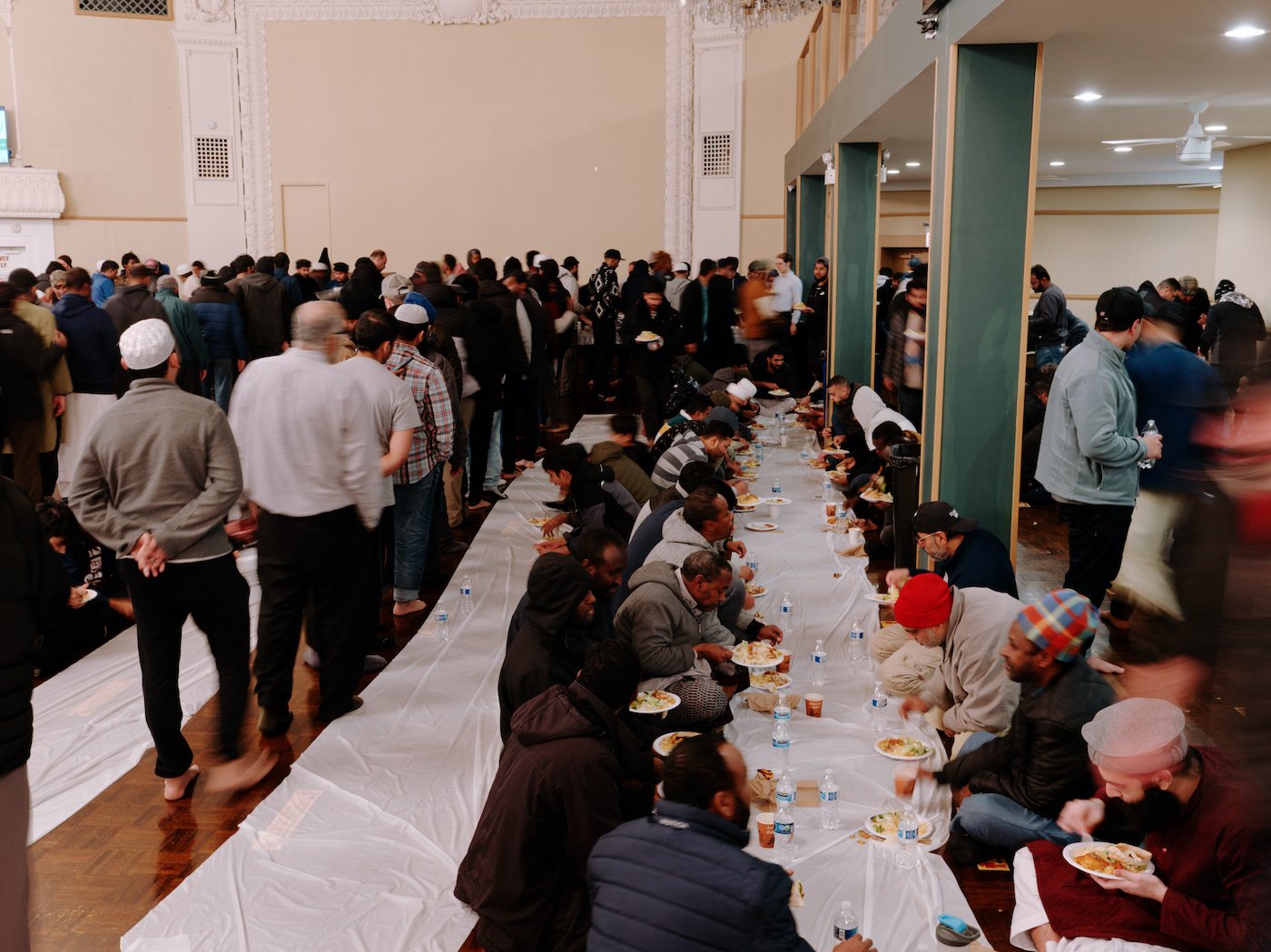 Men sit on the floor and eat from plates on a line of plastic while other men line up to get food