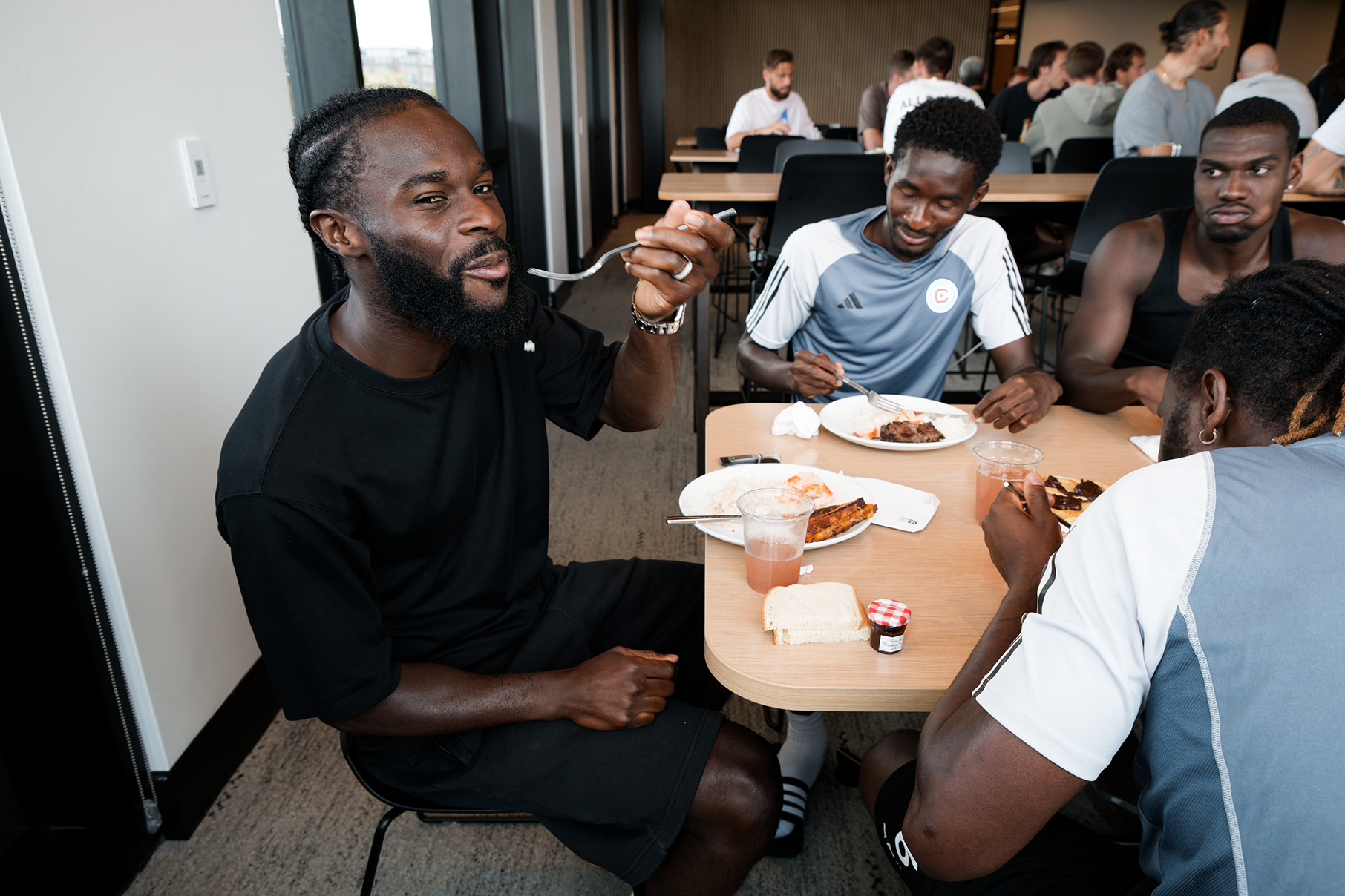 A man smiles as he raises food to his mouth at a cafeteria table with other people