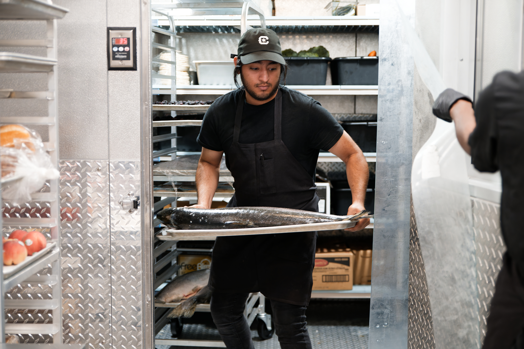 A man carries a whole fish on a tray out of a walk-in fridge