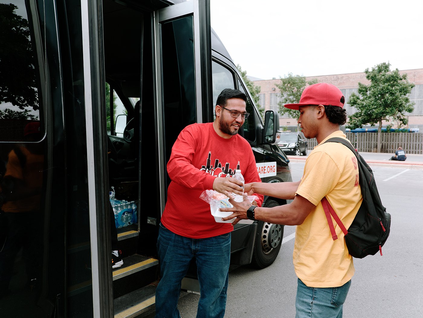 A man in a red shirt smiles and hands over a meal and water to another man