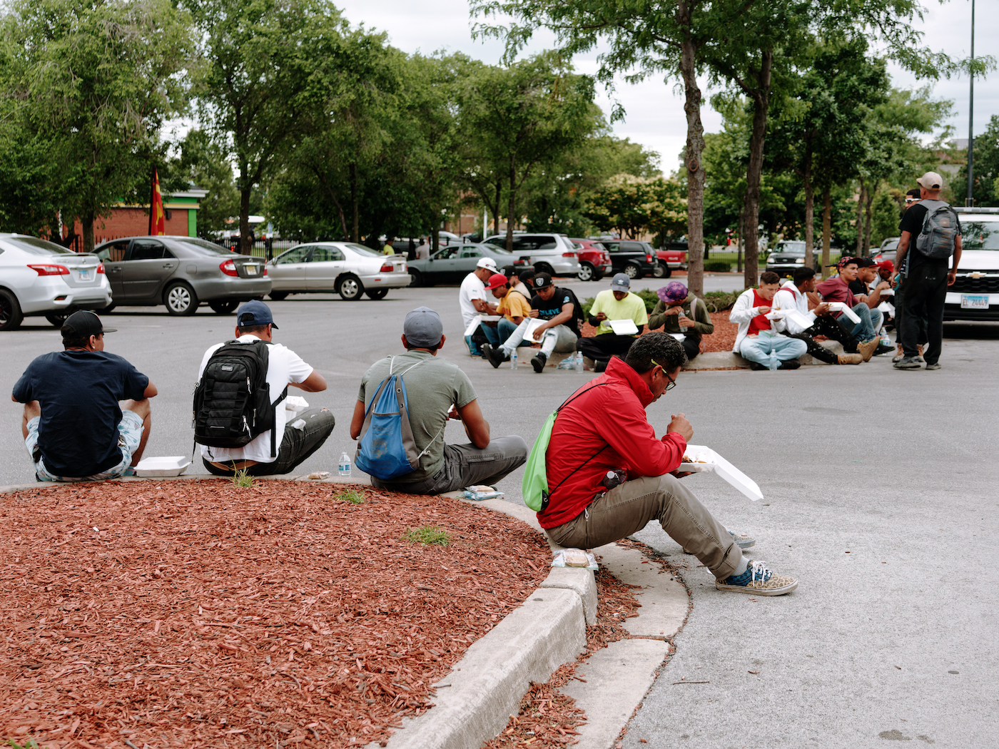 Men seated in a parking lot eat meals