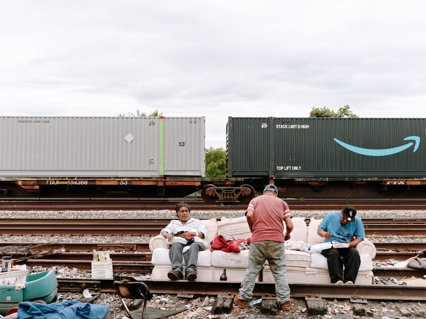 Two men sit on a sofa next to railroad tracks and eat while another stands in front of them