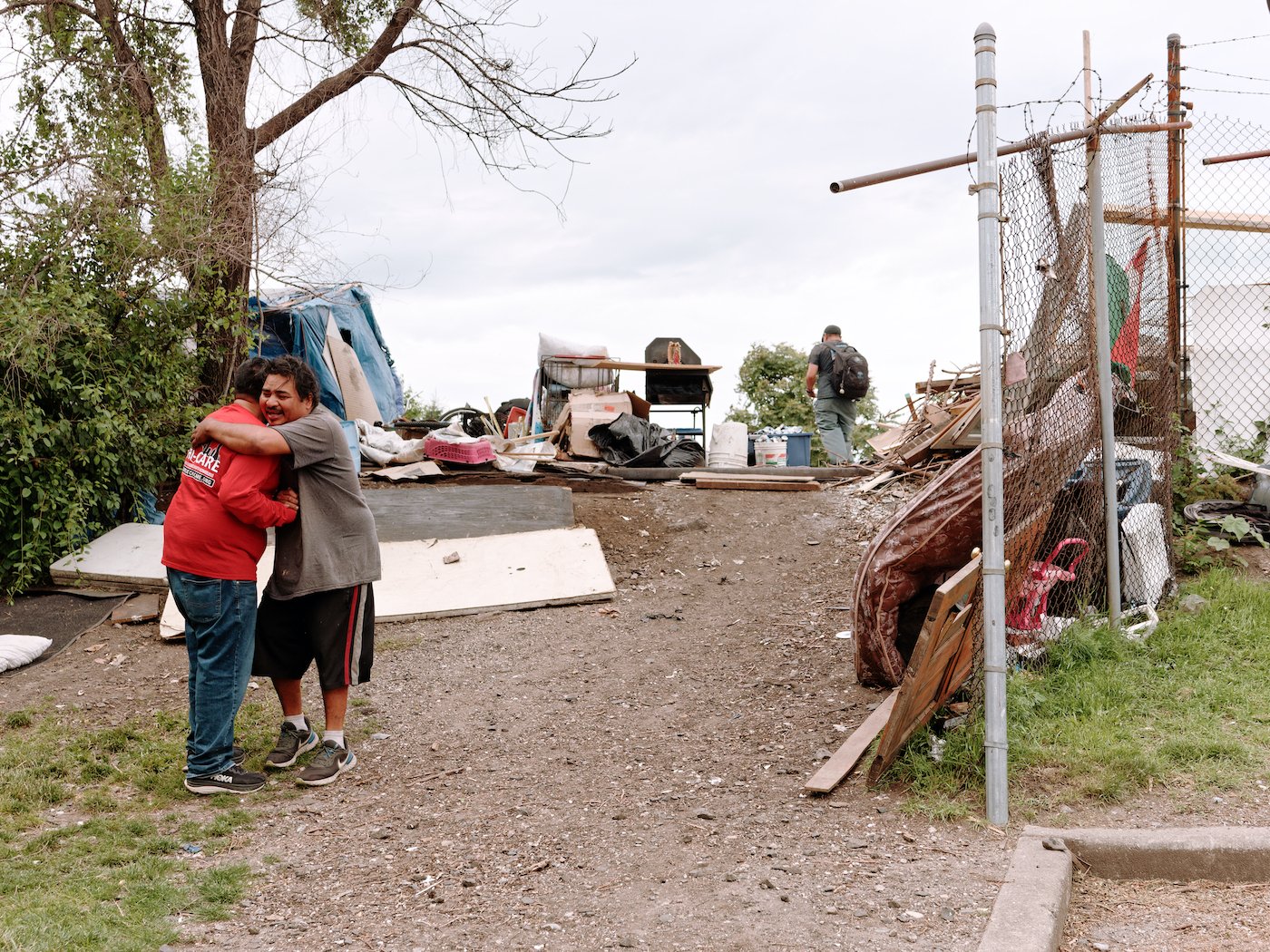 Two men greet each other with a hug on a gravel slope in front of a homeless encampment