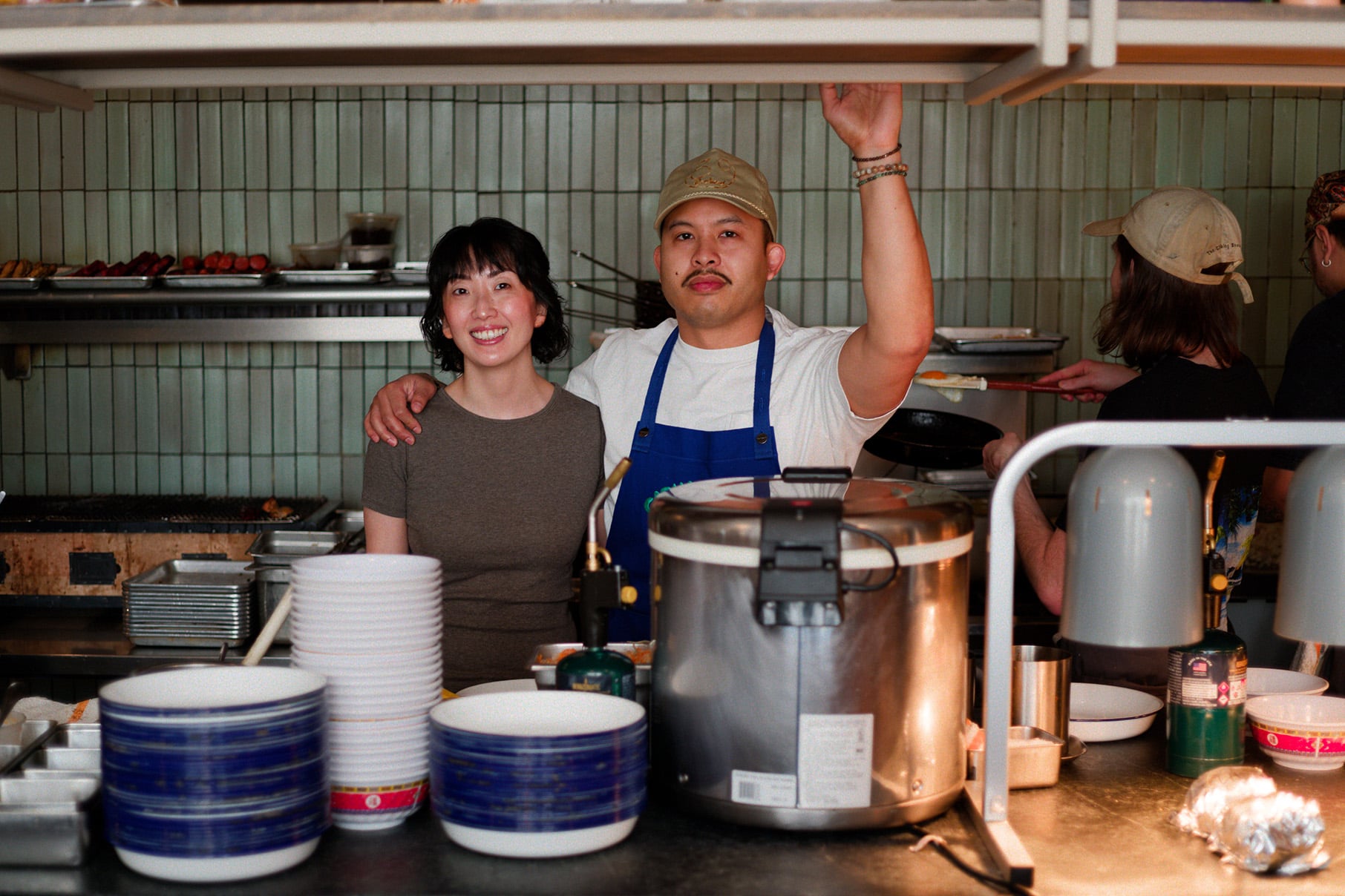 Genie Kwon and Tim Flores stand behind a counter stacked with plates and a rice warmer