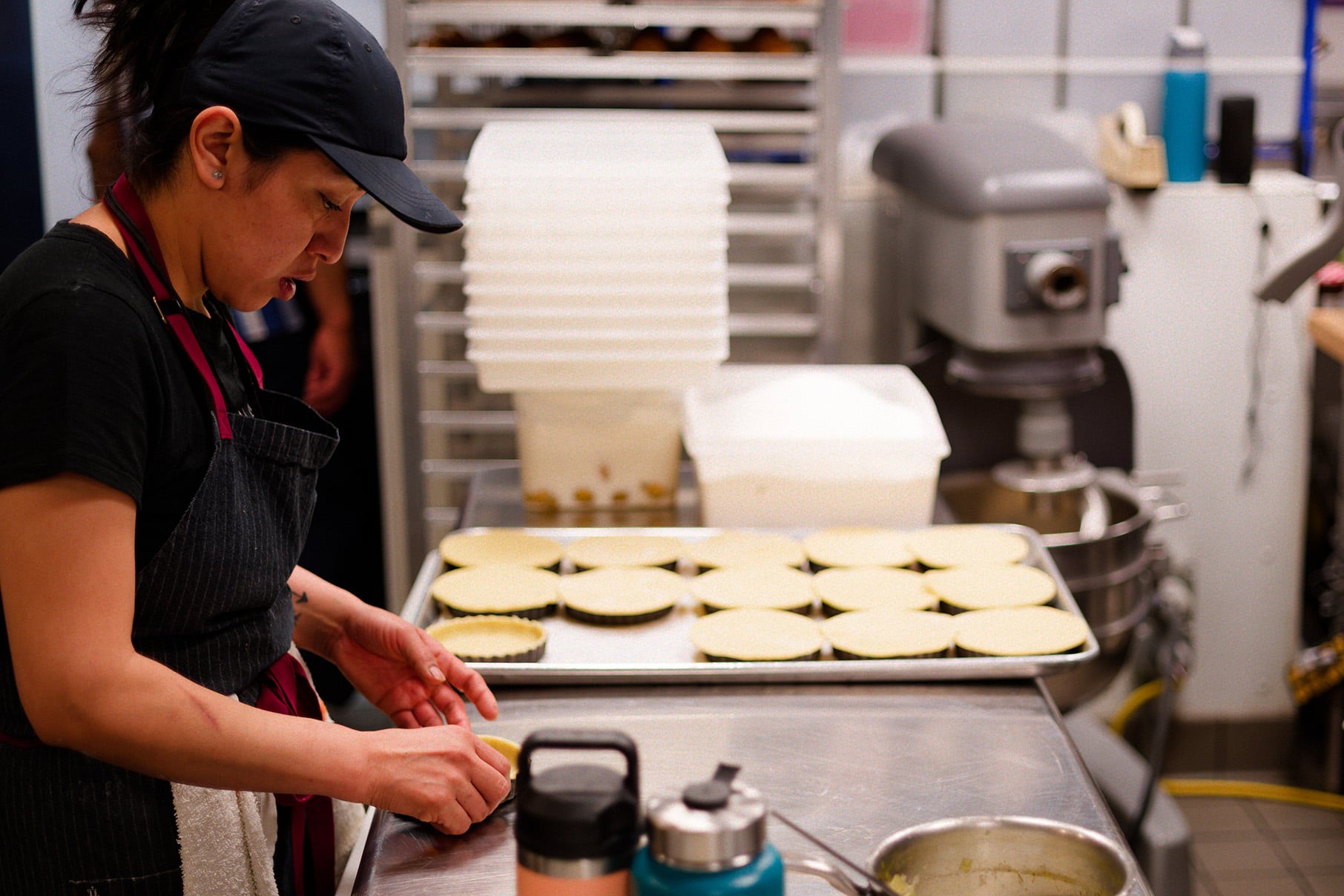 Rows of tart crusts stand next to a woman working on one