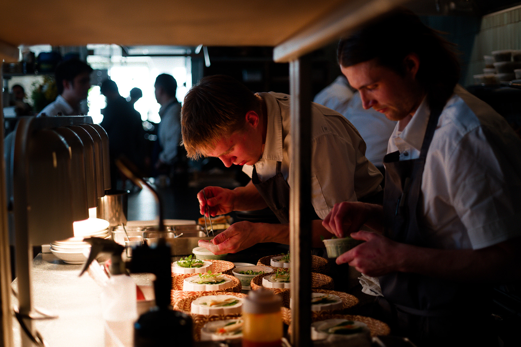 A man in an apron bends over a plate with tweezers in a long open restaurant kitchen