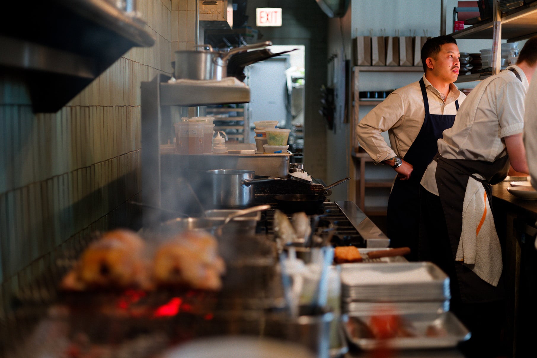 A grill is blurred in the foreground while Tim Flores stands in an apron near another person in the background