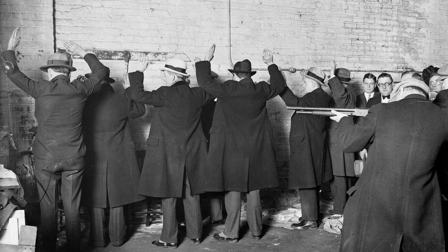 A group of men in coats and hats with their hands up against a wall as another man holds a gun pointed at them.