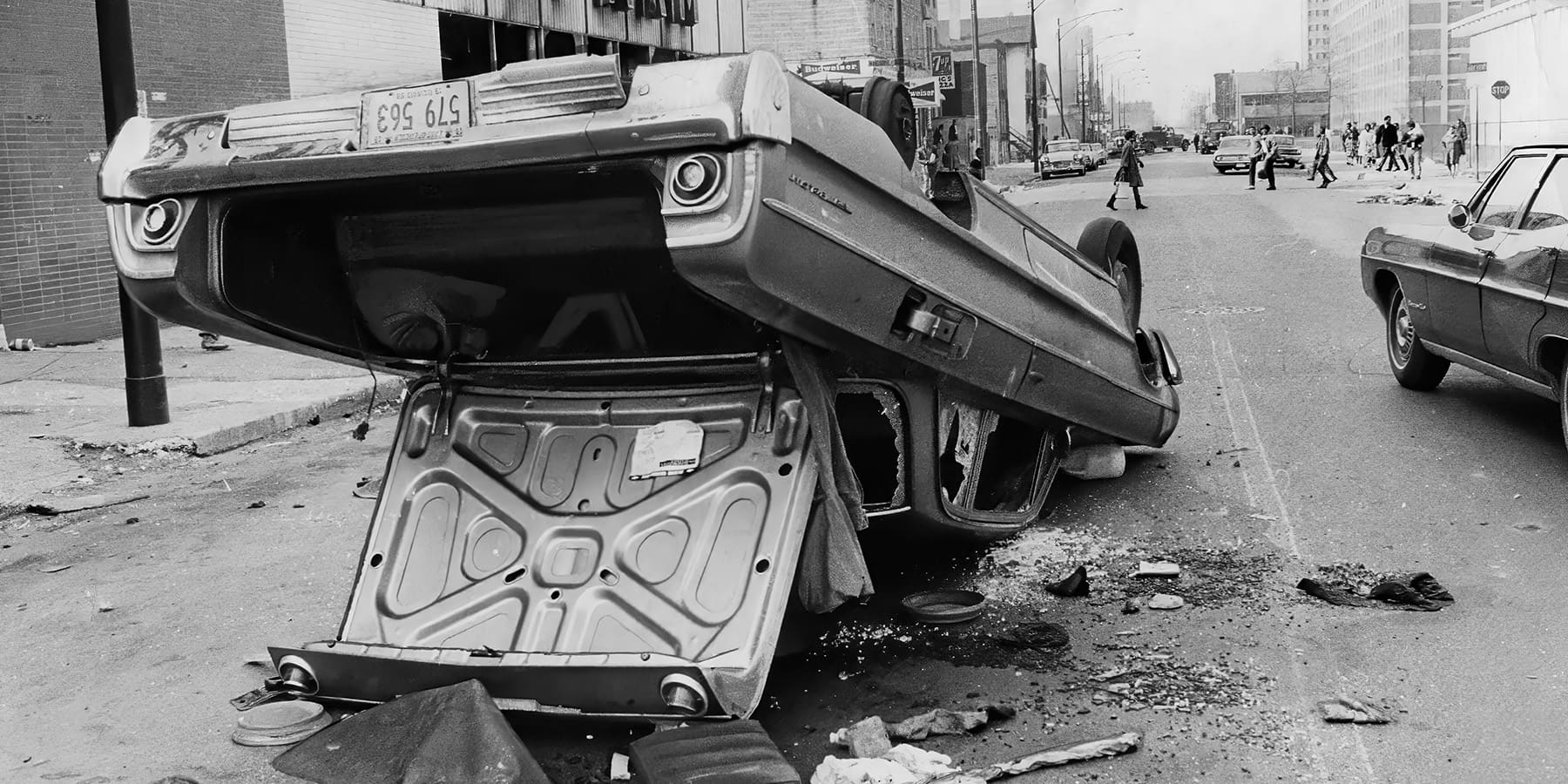 A black-and-white image of a car overturned on a city street. The underside of the car is visible, with debris scattered around it.