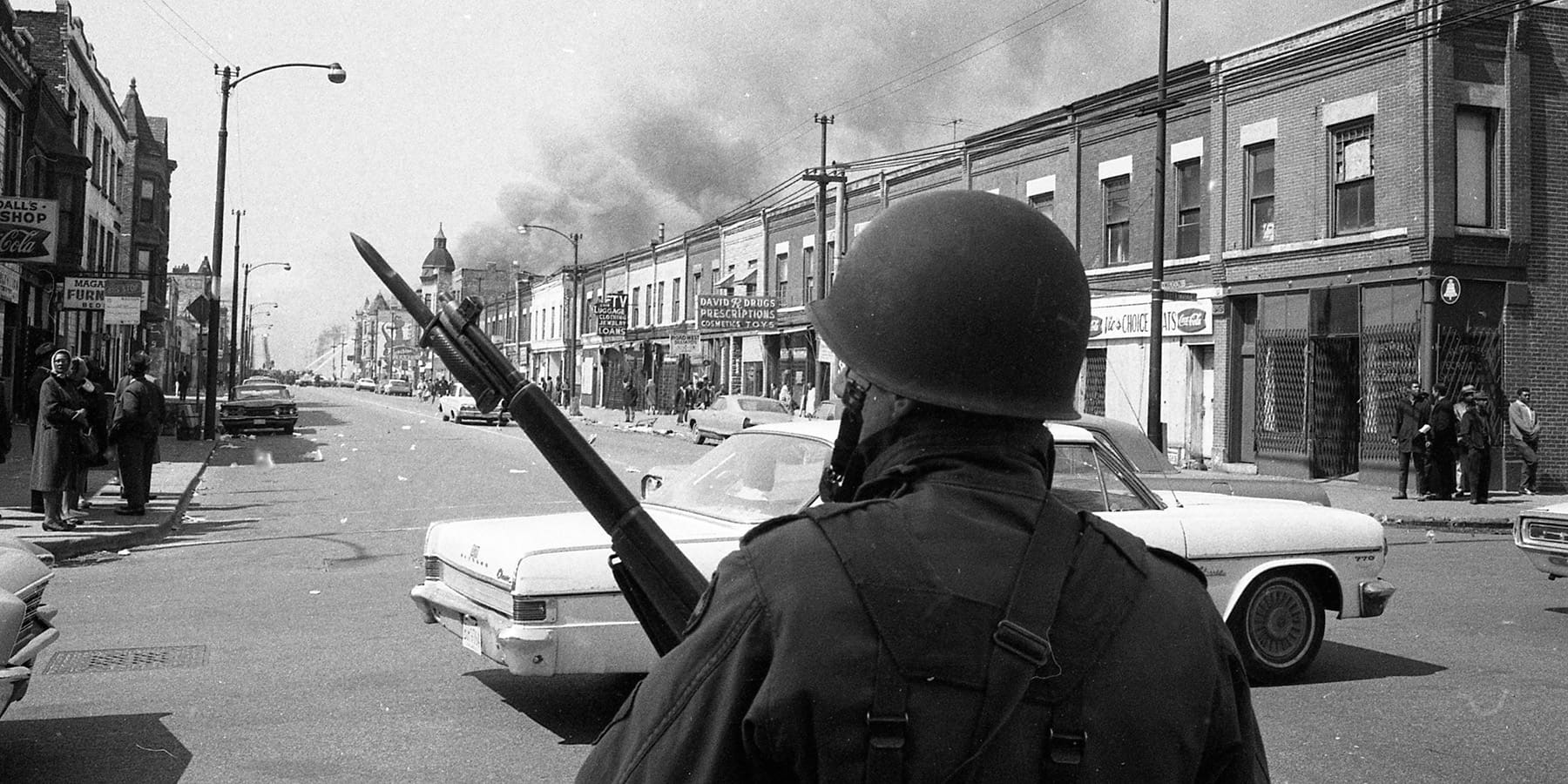 A soldier in uniform and helmet, holding a rifle, stands in the middle of a city street. Buildings are on either side, and smoke rises in the background.