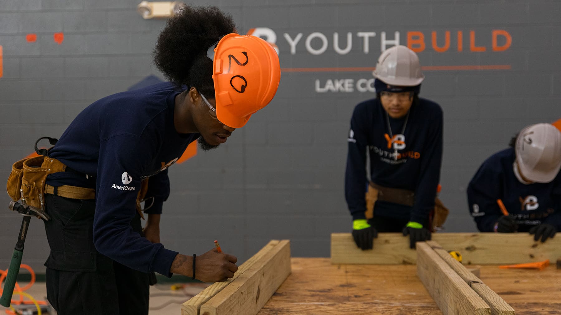 A young African American male wearing a hard hat and toolbelt marks a piece of lumber on a workbench
