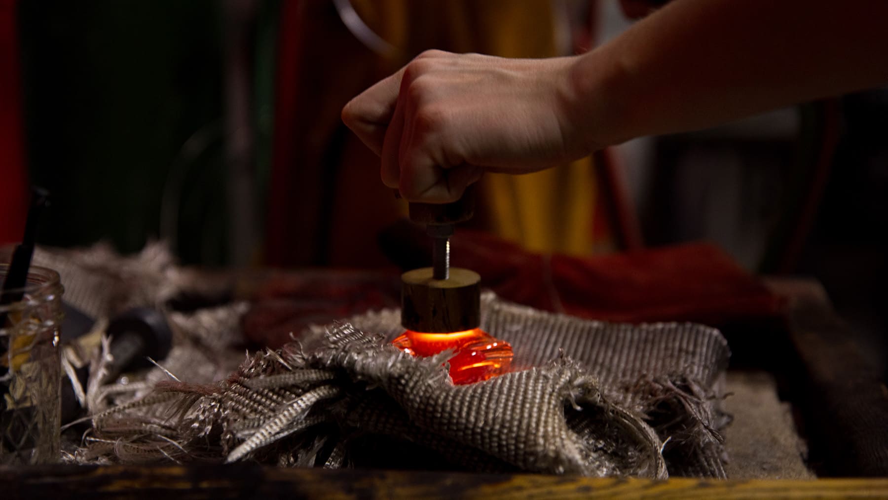 A hand presses down a stamp onto orange glowing molten glass