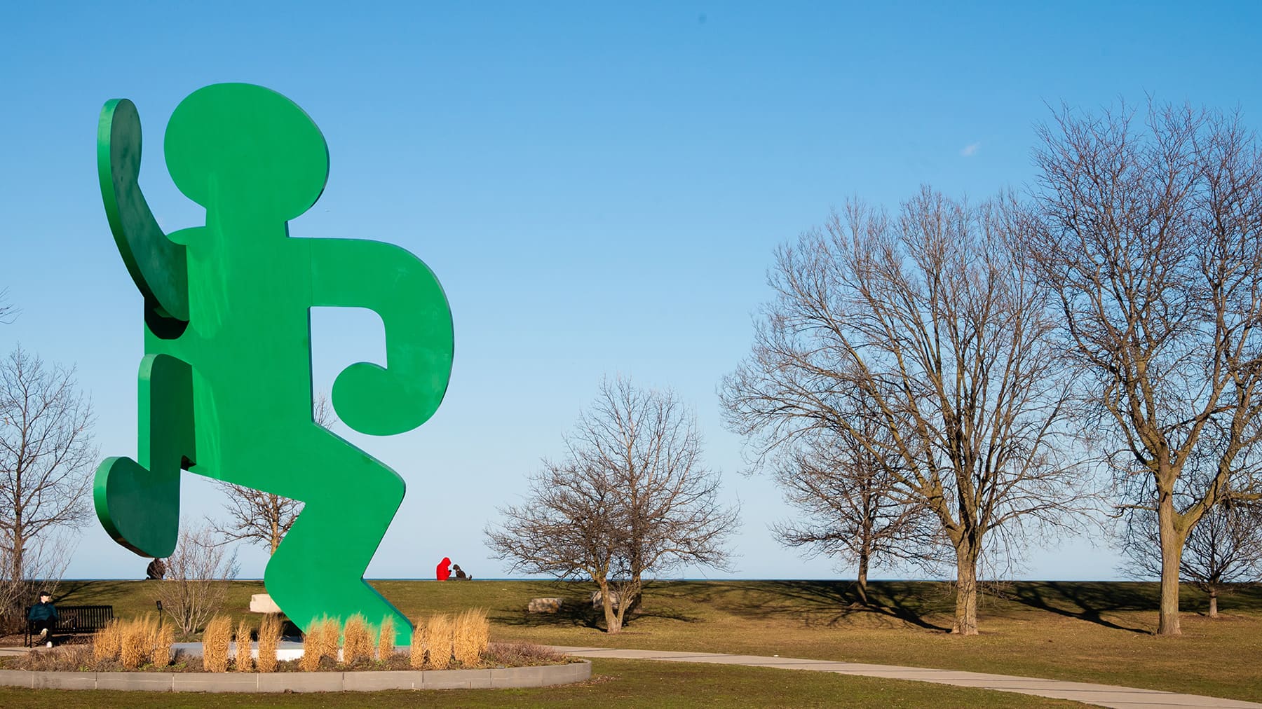 A stature of a green stylized human figure stands with one arm and leg raised with trees and a blue sky in the background.