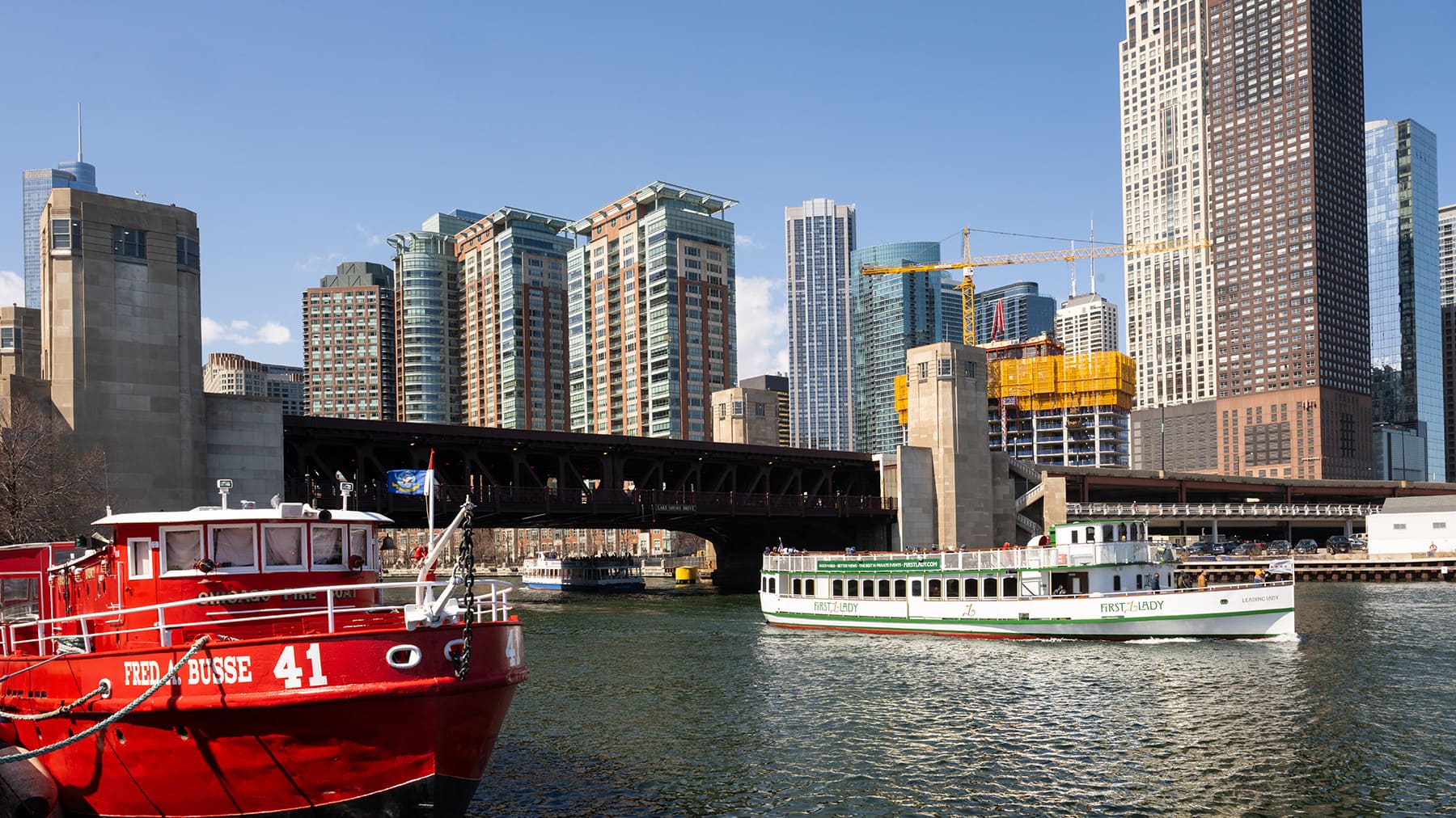 A docked Chicago Fire Boat and a tour boat are on the Chicago River with an iron bridge and highrises in the background.