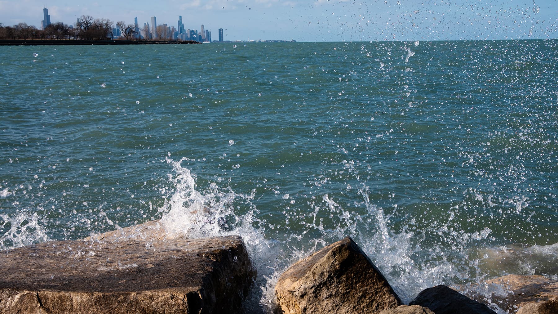 A wave crashes against the shoreline rocks with the Chicago skyline in the background.