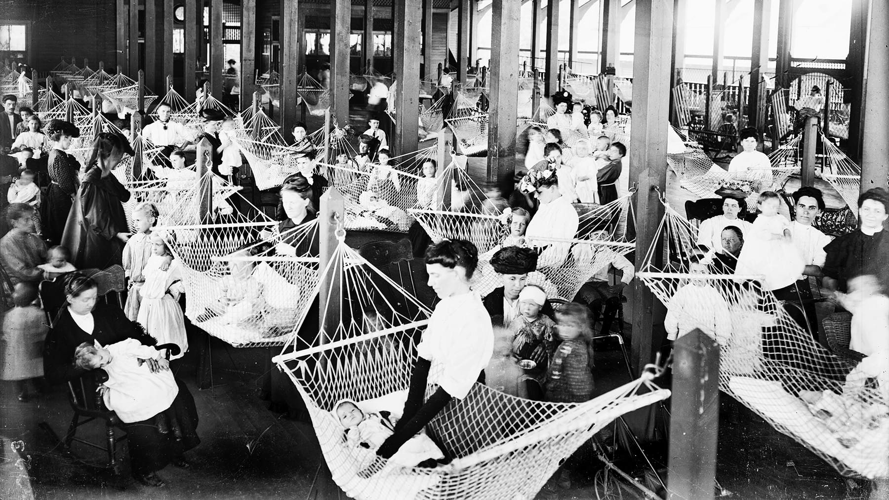 A historical photo showing a large room filled with hammocks, young children and caregivers.