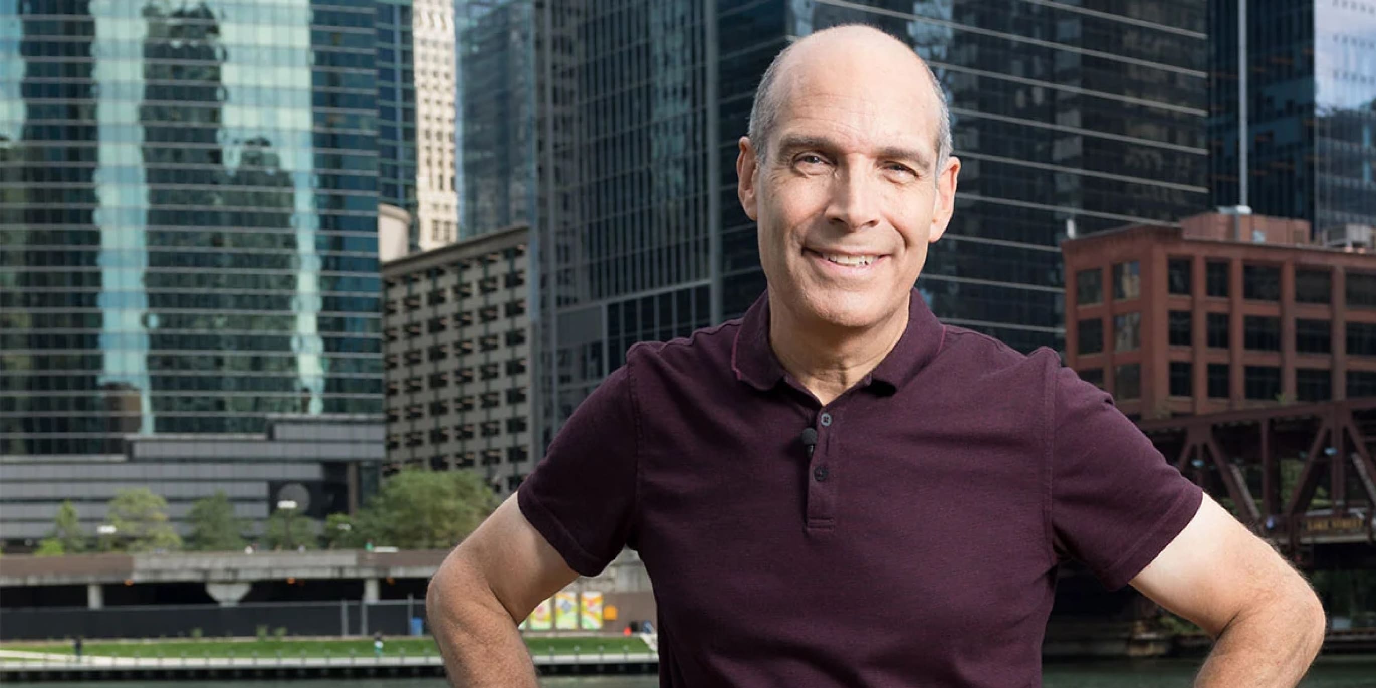 Geoffrey Baer stands on the river bank with buildings in the background.