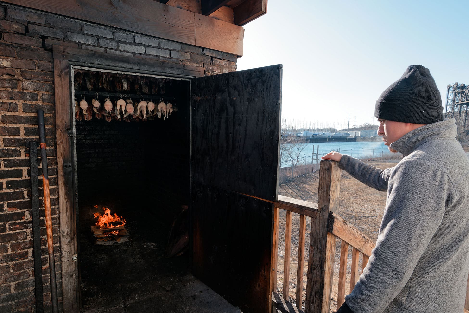 A worker stands outside the smokehouse as fish hangs above the burning logs.