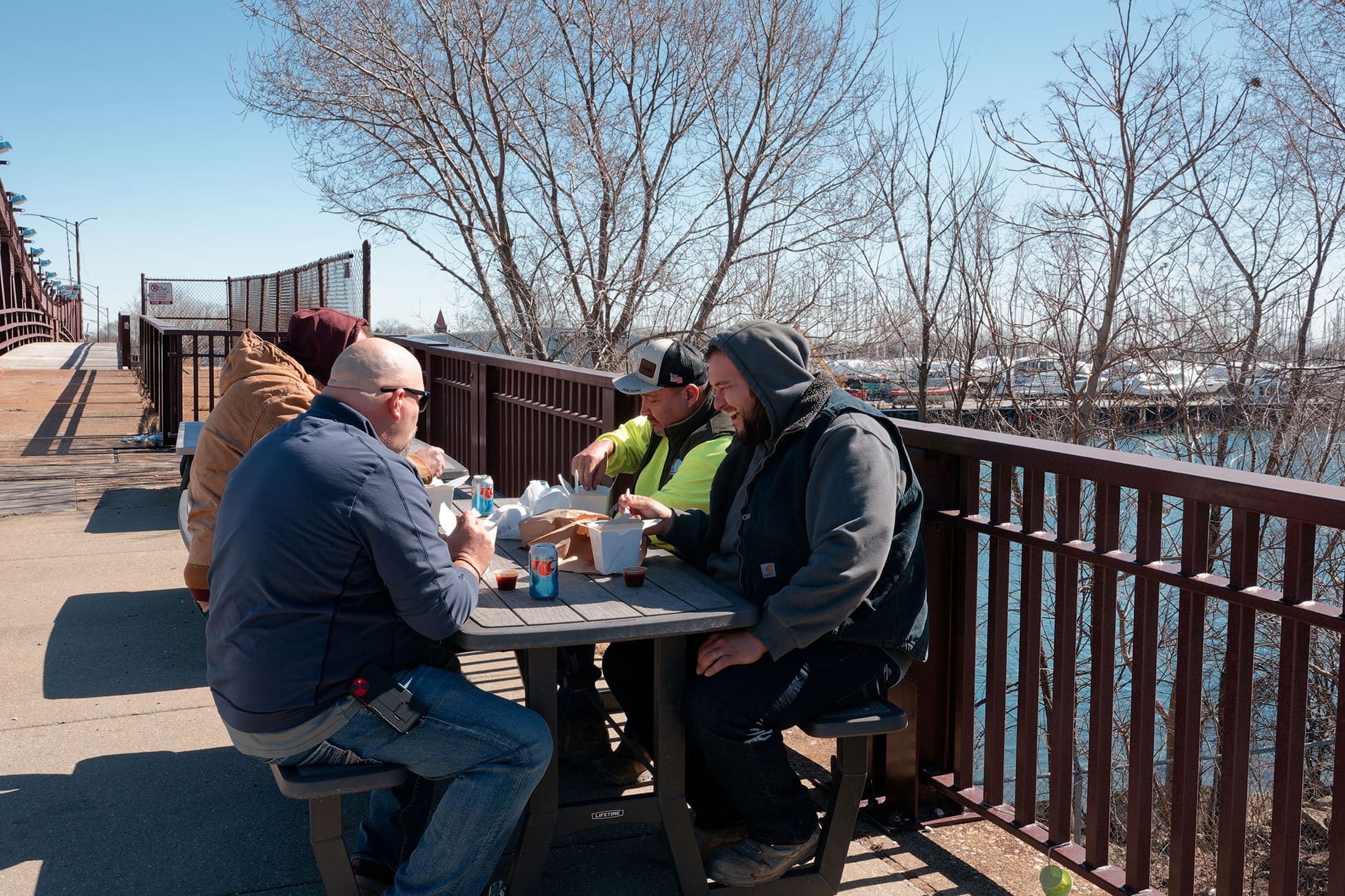 Four men sit at a picnic table outside to eat their meals.