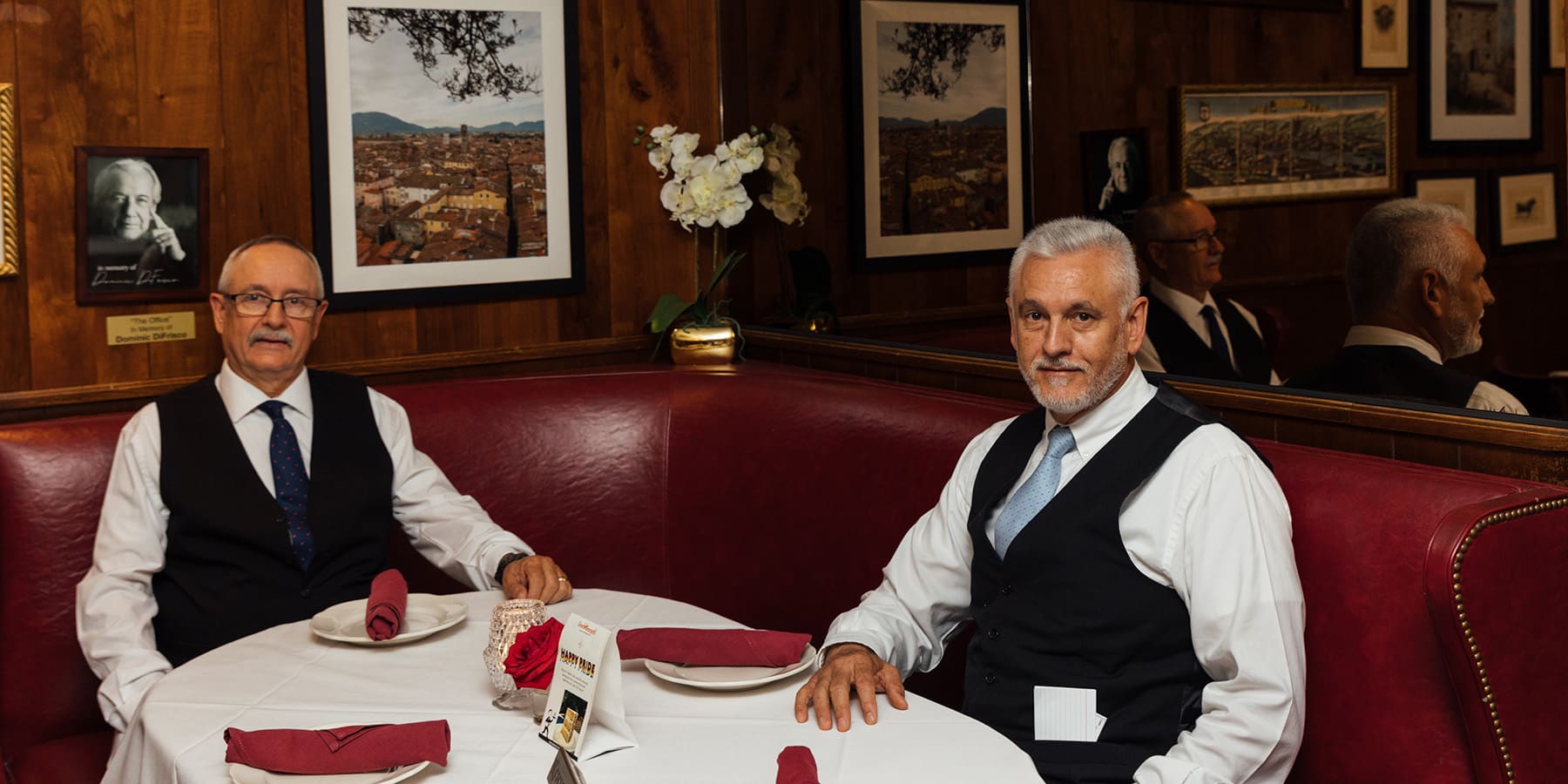 Two men in white shirts, ties and vests sit in a corner red-leather booth of a restaurant