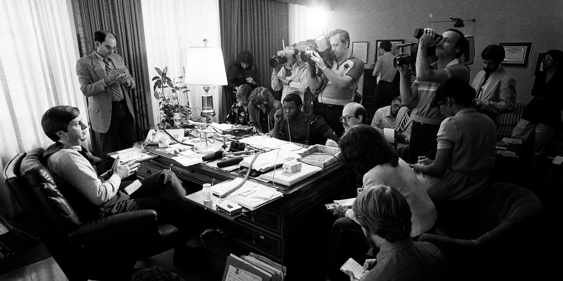 Tyrone Fahner sits at his desk surrounded by reporters and cameramen