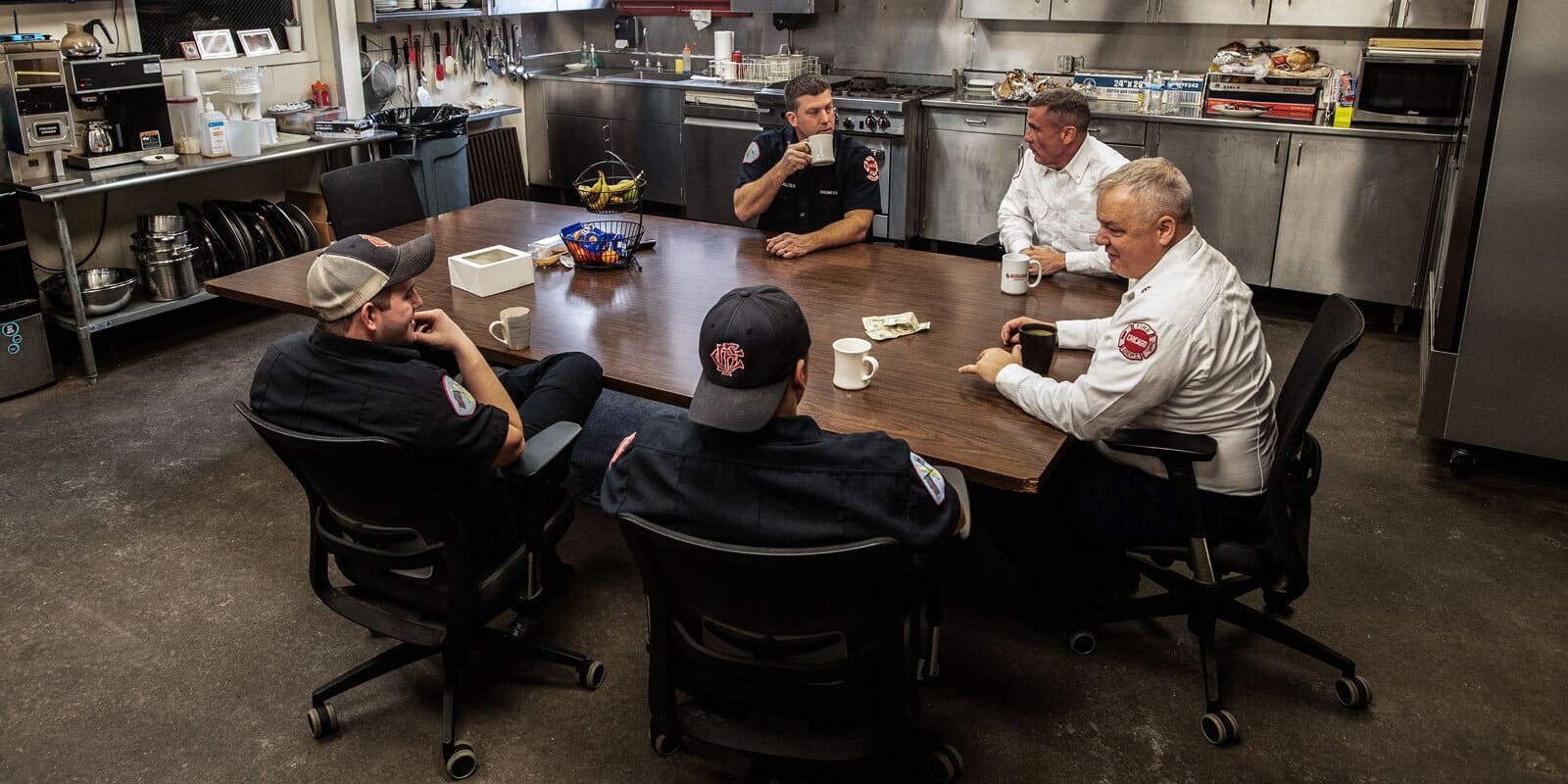 Five firemen sit around the kitchen table talking and drinking coffee