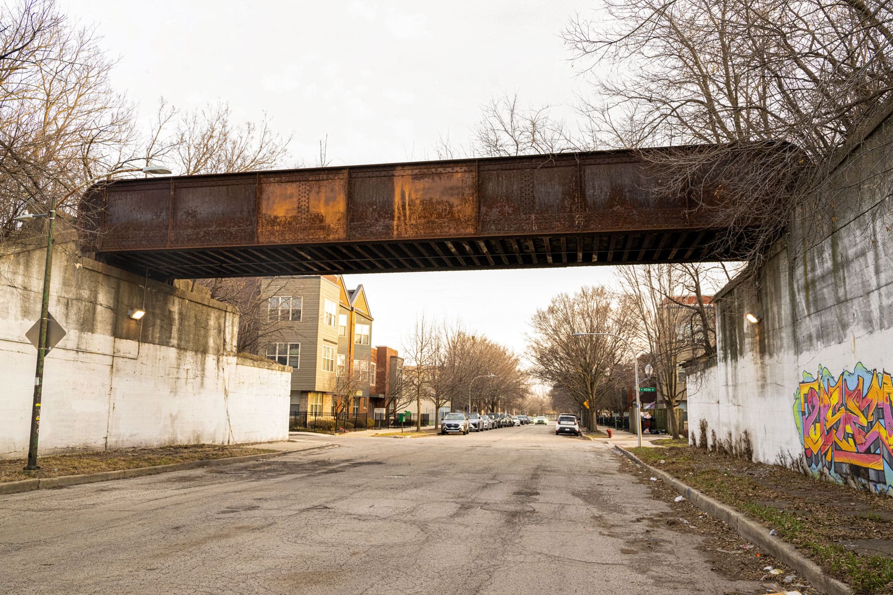 Urban scene with a rusty iron bridge spanning a quiet street, flanked by winter trees and colorful graffiti on the wall, under an overcast sky.