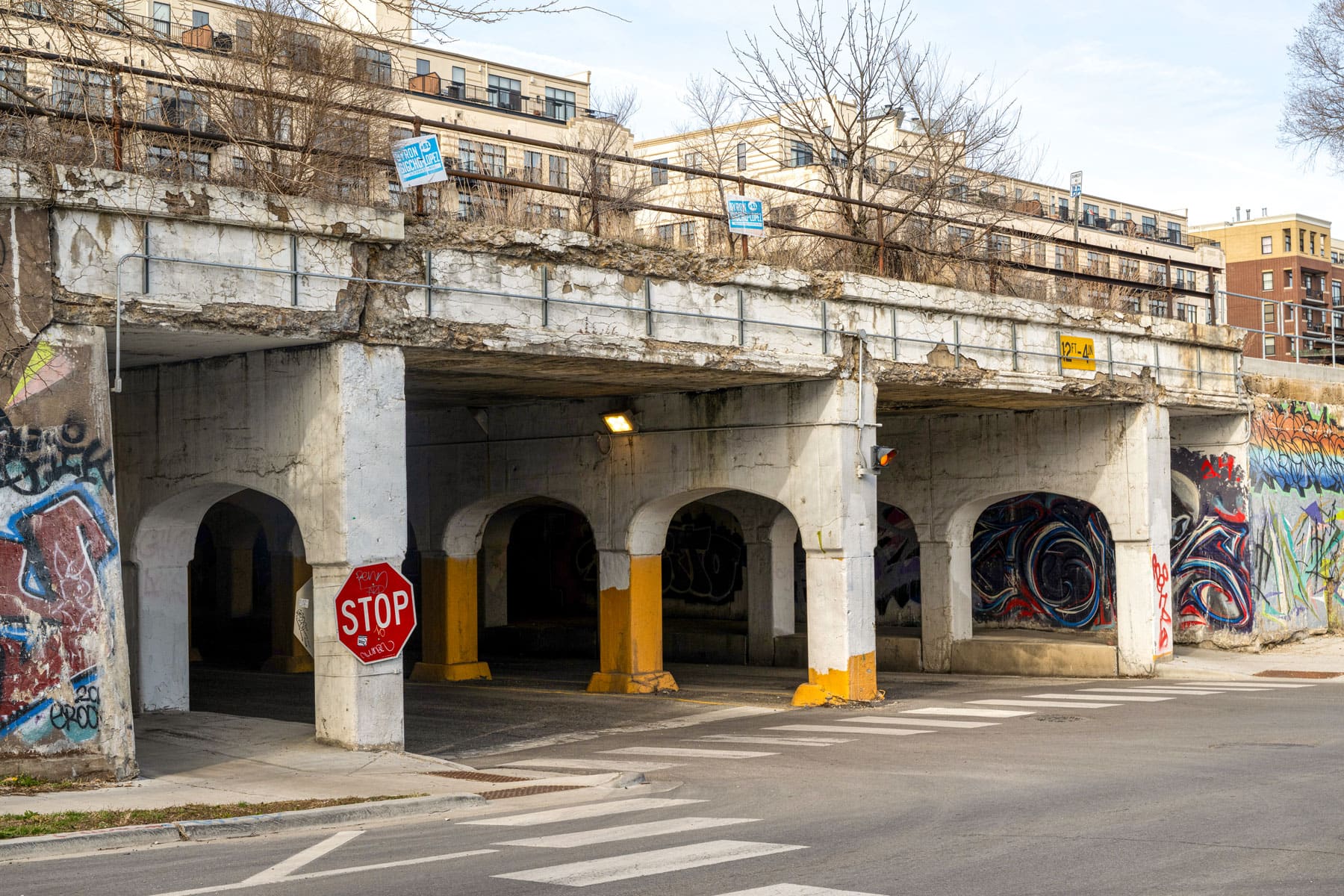 Weathered concrete overpass with graffiti, a stop sign, and urban buildings in the background. The scene conveys an urban, gritty ambiance.