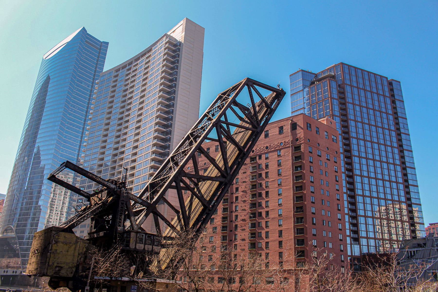 A rusted railway drawbridge in the foreground contrasts with modern glass and brick skyscrapers under a clear blue sky, blending old and new architecture.