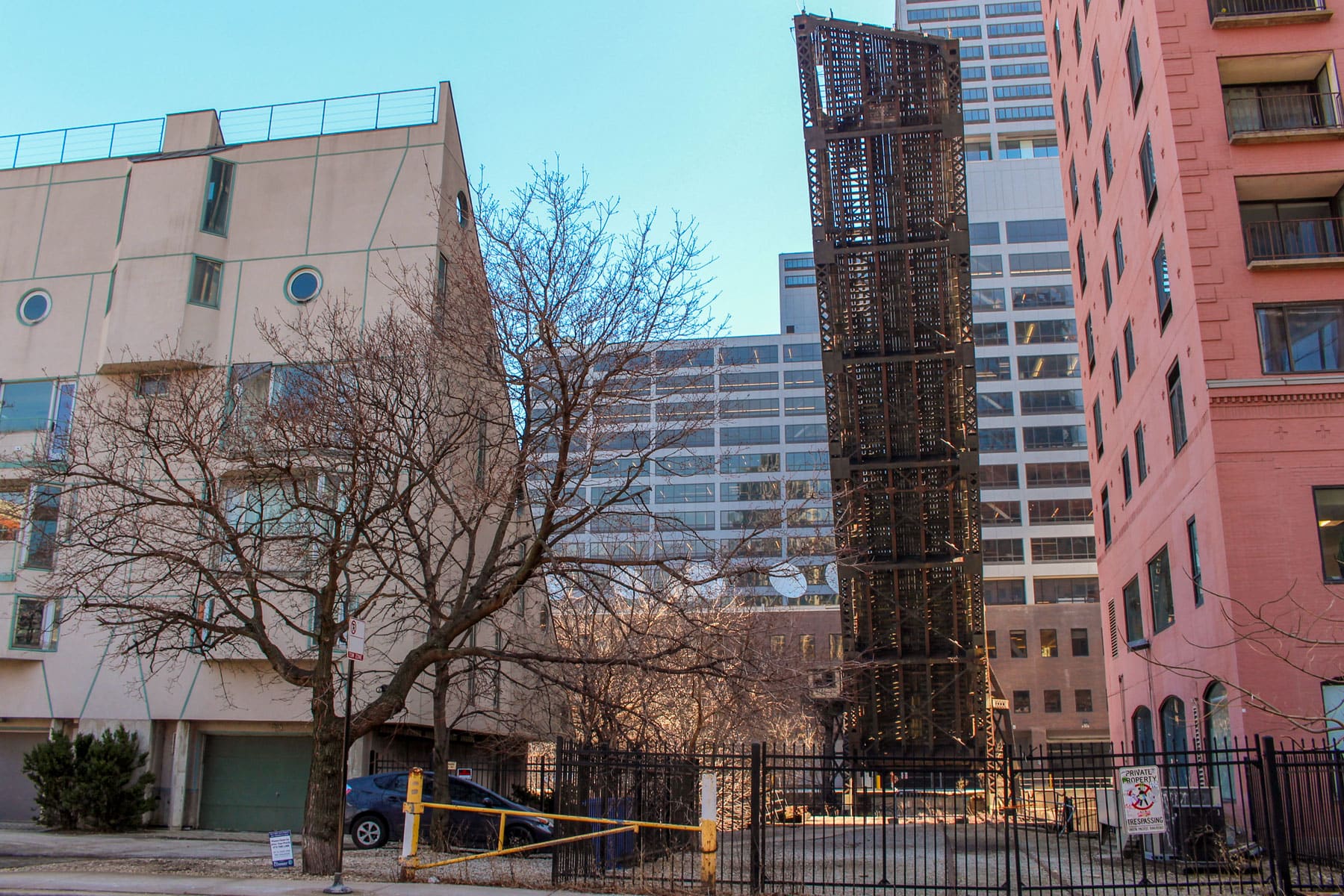 Downtown scene with tall, rusty bridge lifted vertically between modern buildings. Bare trees, a parked car, and blue sky add to the urban atmosphere.