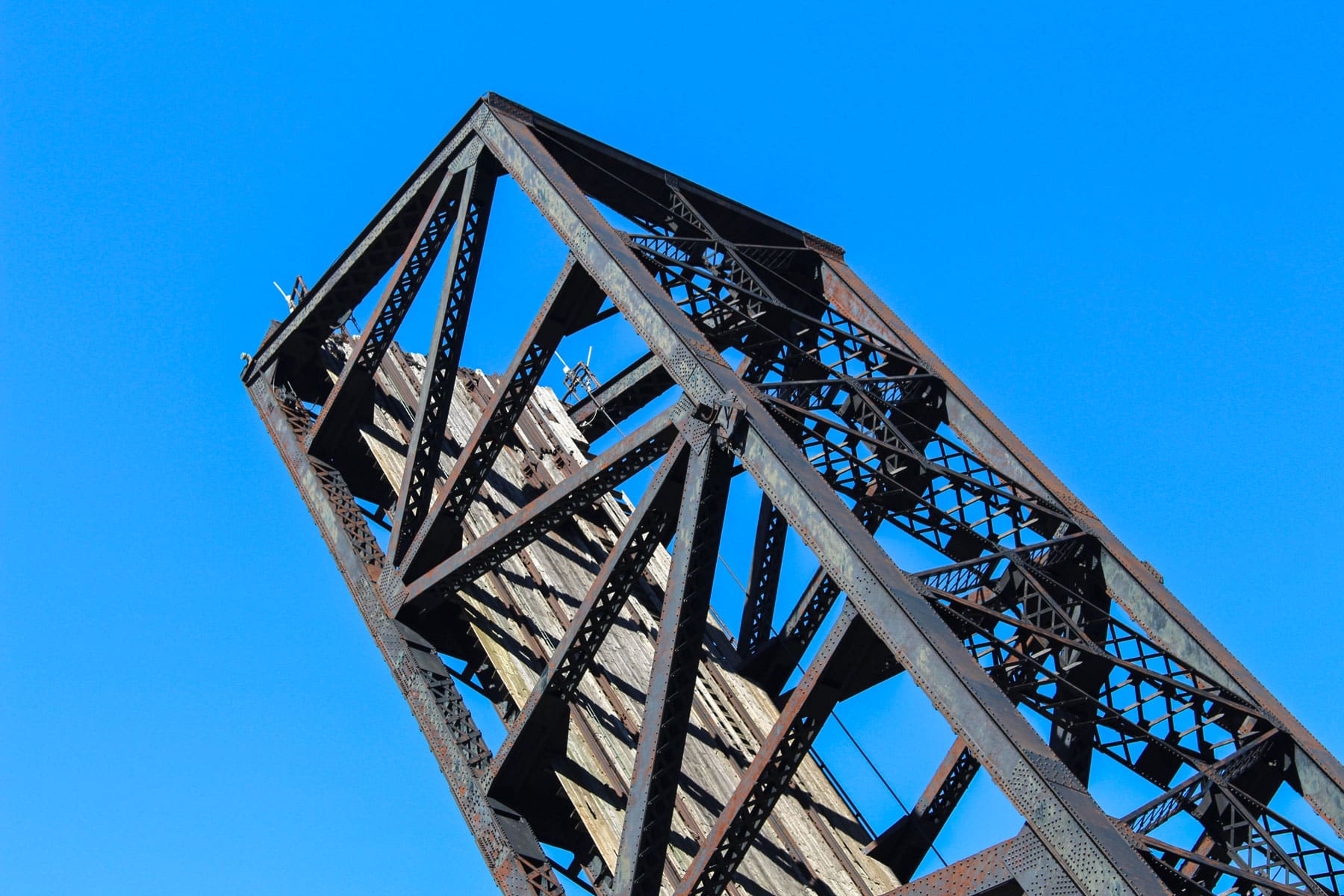Rusty, weathered drawbridge seen from below against a clear blue sky. The steel beams form intricate patterns, conveying a sense of abandonment.