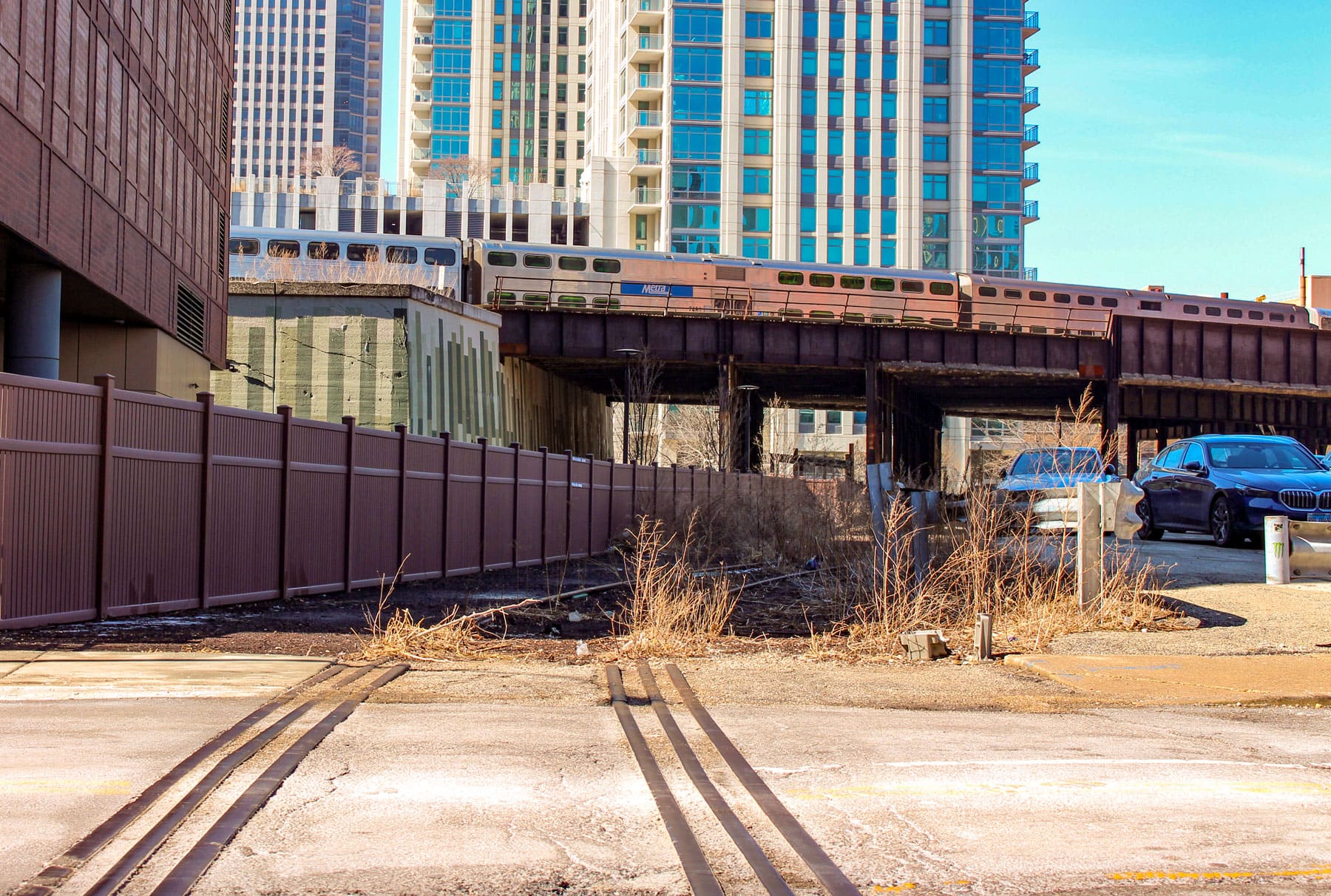 Urban scene with a train crossing a rusty bridge against tall buildings. Foreground shows overgrown weeds and parked cars, conveying an industrial vibe.