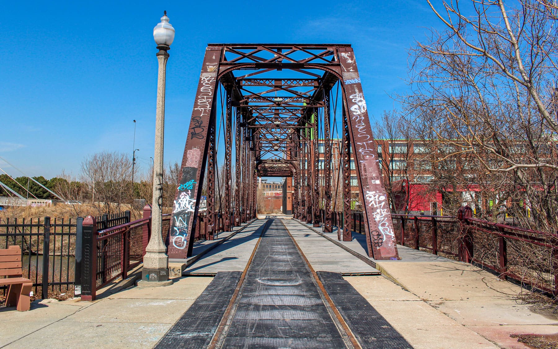 Rustic metal truss bridge with graffiti spans a pathway. Bare trees and urban buildings line the background under a clear blue sky.