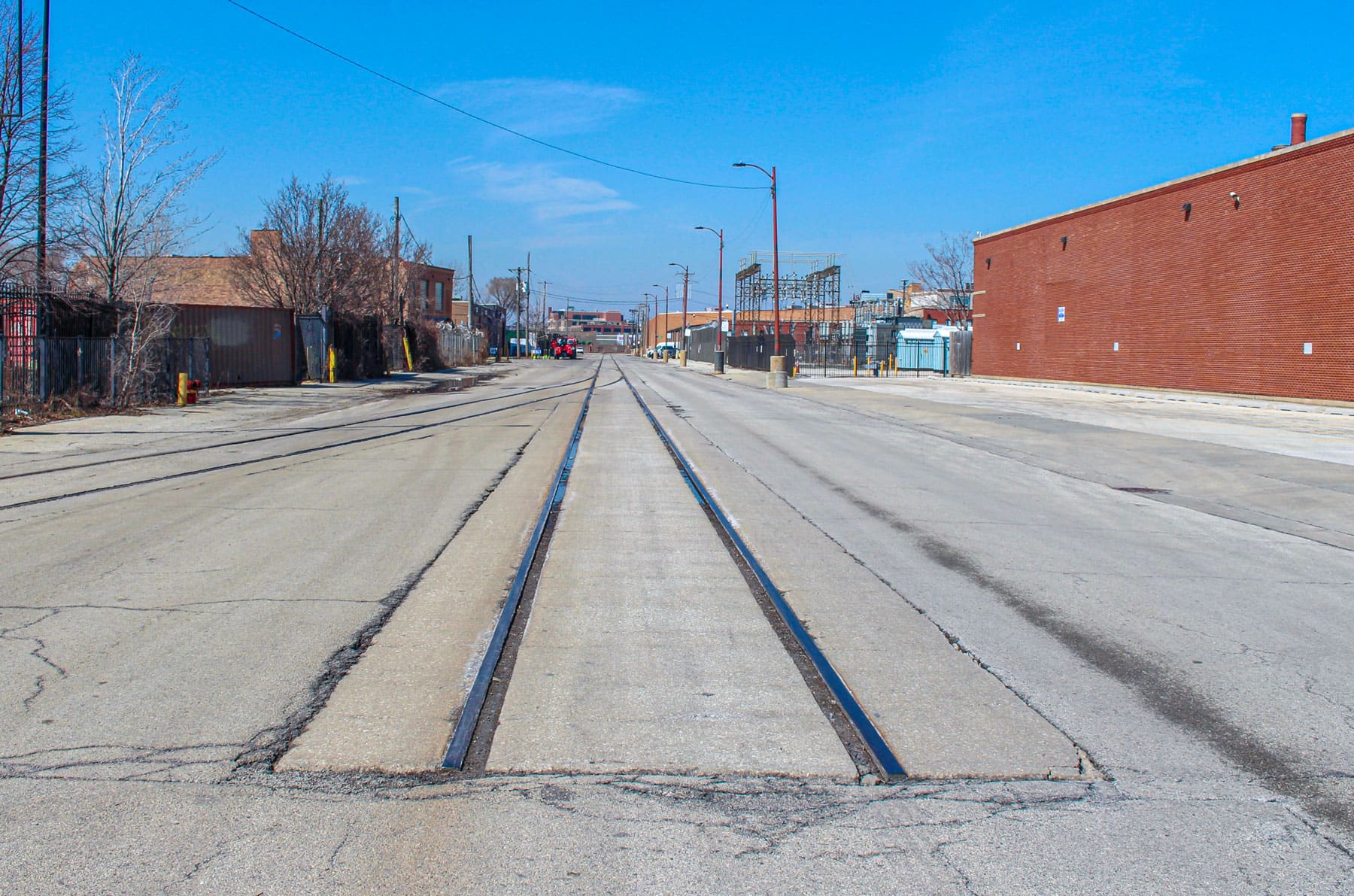 Empty urban street with old train tracks embedded in the pavement, flanked by industrial buildings and bare trees under a clear blue sky.