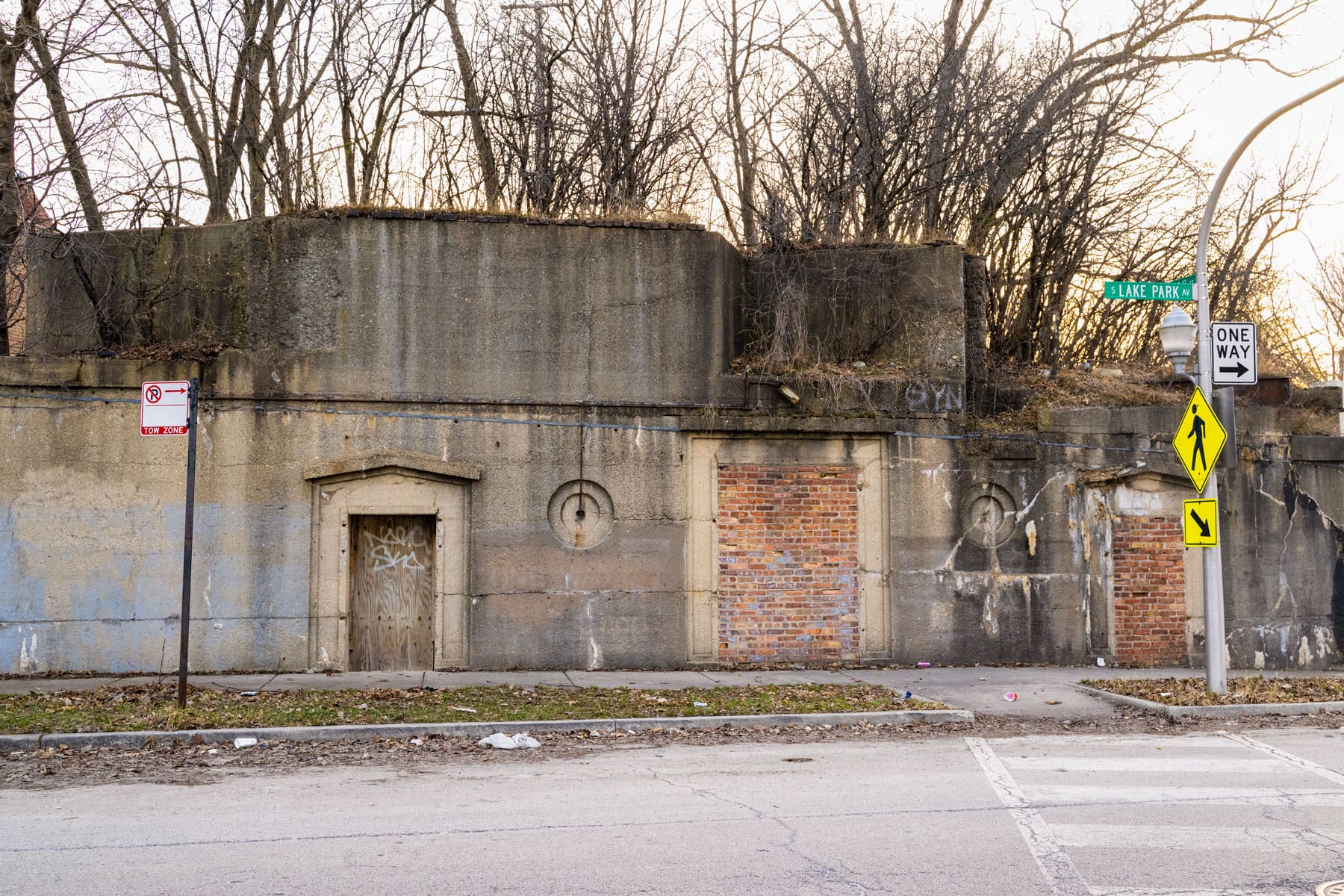 An old, graffiti-covered concrete and brick wall with a wooden door sits under leafless trees.