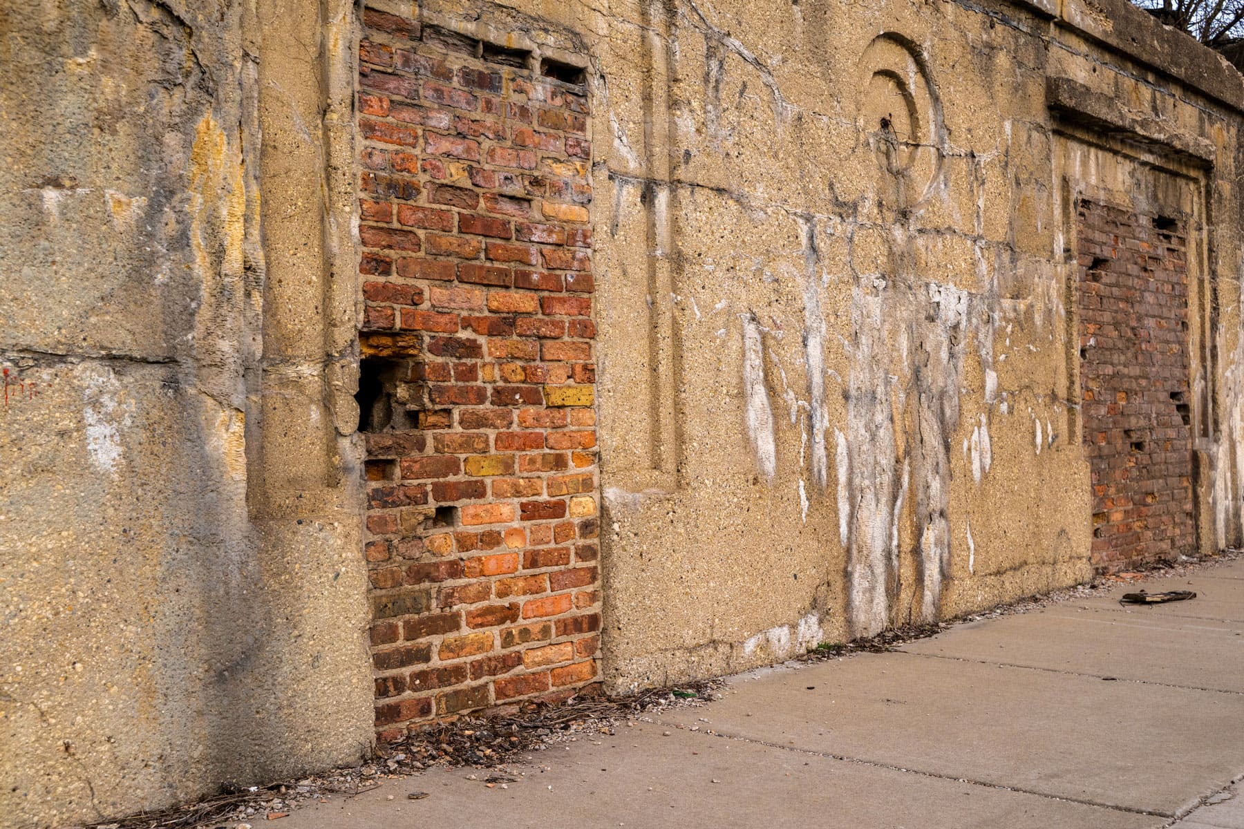A weathered brick wall with two bricked-up doorways and a faded plaster facade. The surface is stained, evoking a sense of decay and urban history.
