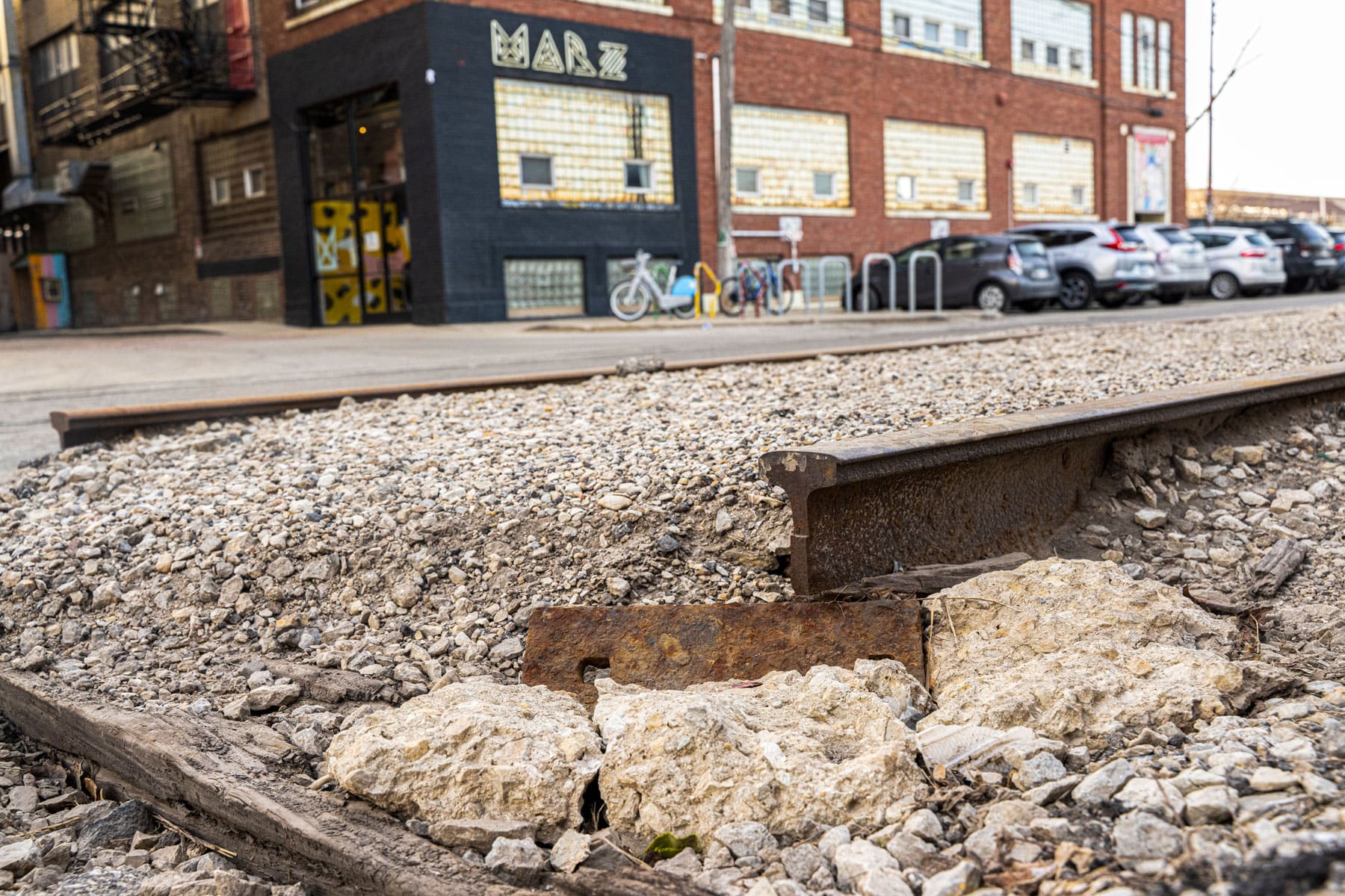 Close-up of an old railway track embedded in gravel, with a background of a brick building and parked cars. The scene conveys urban decay.