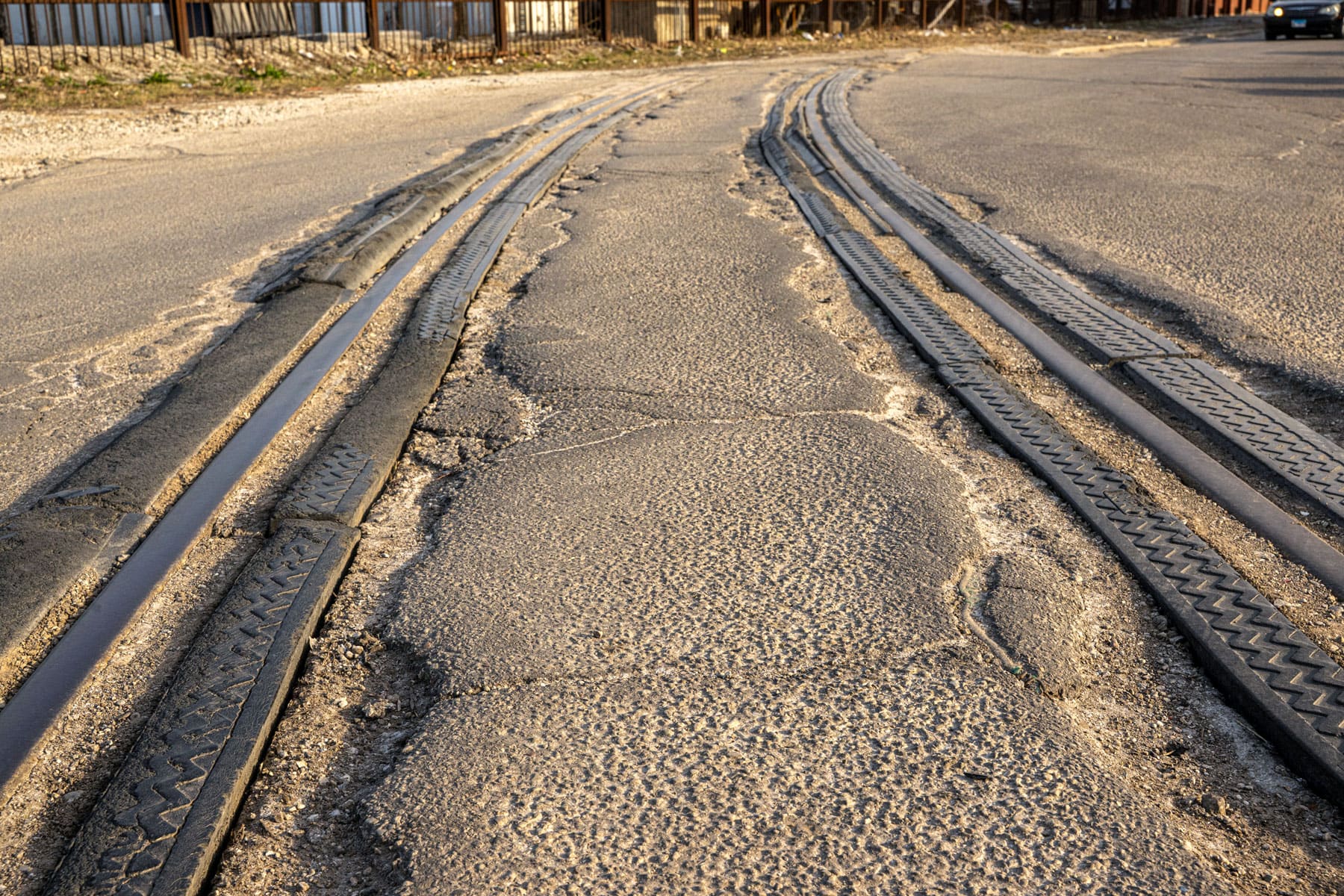 Cracked, worn tram tracks on a deserted urban street, surrounded by rough, uneven asphalt. The scene conveys a feeling of neglect and deterioration.