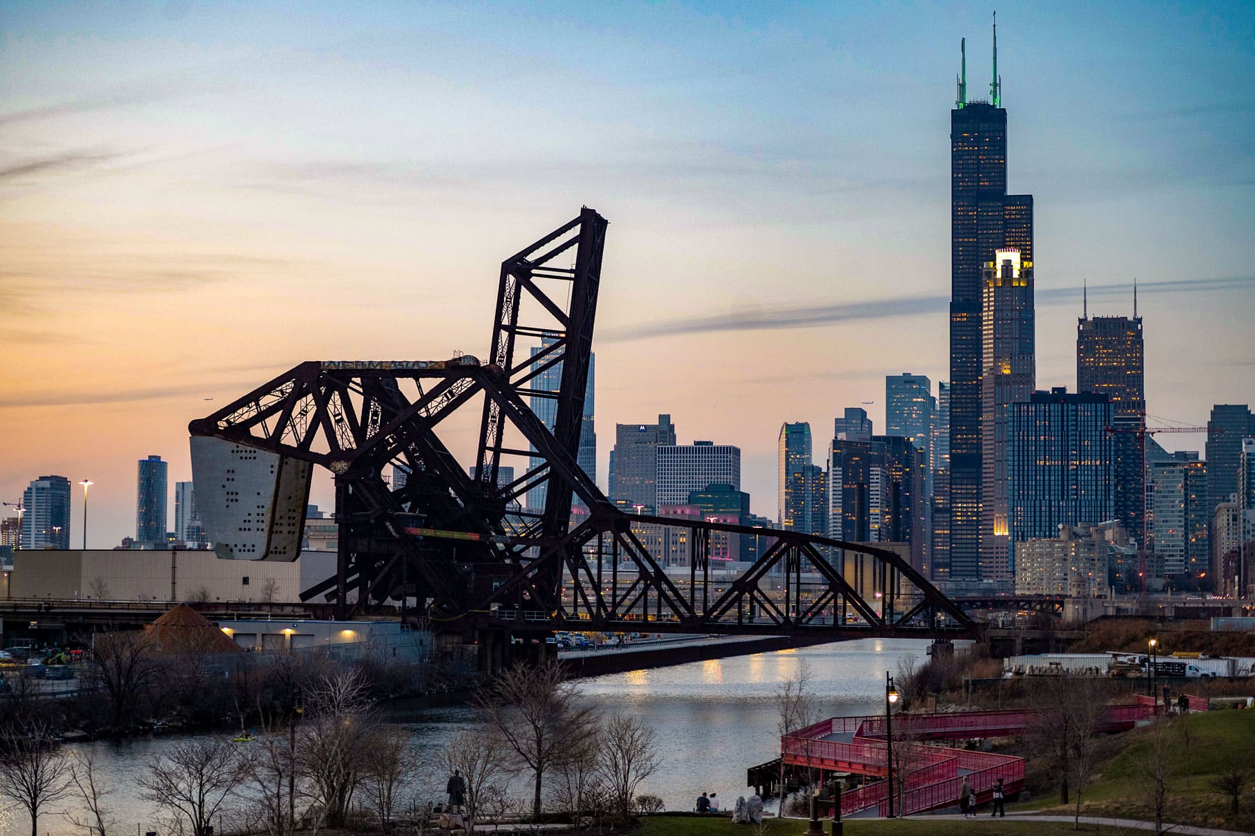 A raised drawbridge spans a river against a city skyline at dusk, with skyscrapers reflecting warm lights. The sky is gradient blue and orange.
