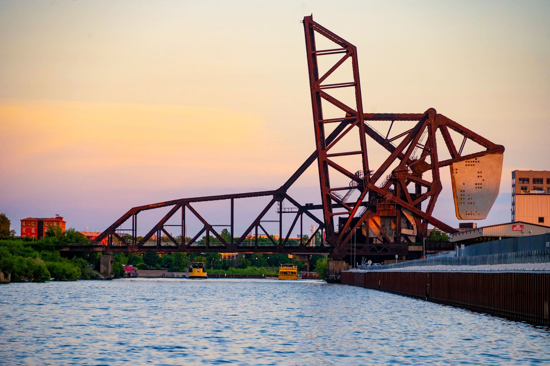 Rusted drawbridge over a calm river with two yellow water taxis beneath. The sky is softly lit by a pastel sunset, adding a serene ambiance.