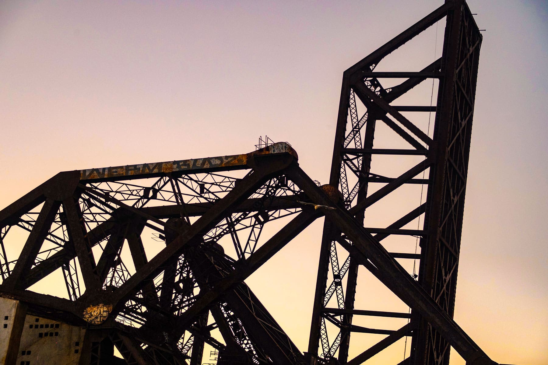 Rusty steel train bridge lifted against a pinkish sky, creating a silhouette. The industrial structure evokes a sense of abandonment and history.