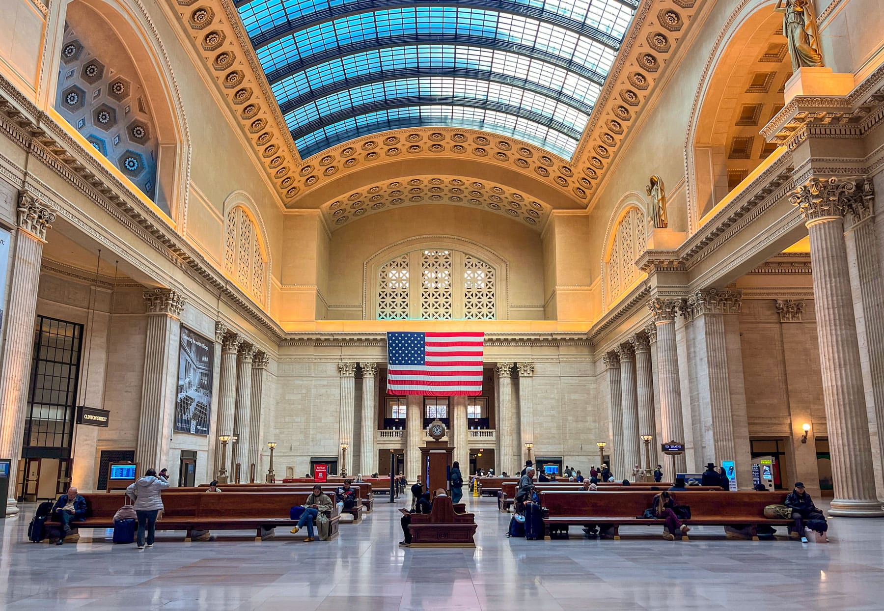 The spacious interior of Union Station with a large American flag hanging under an ornate arched ceiling. People sit on benches, and a clock stands central.