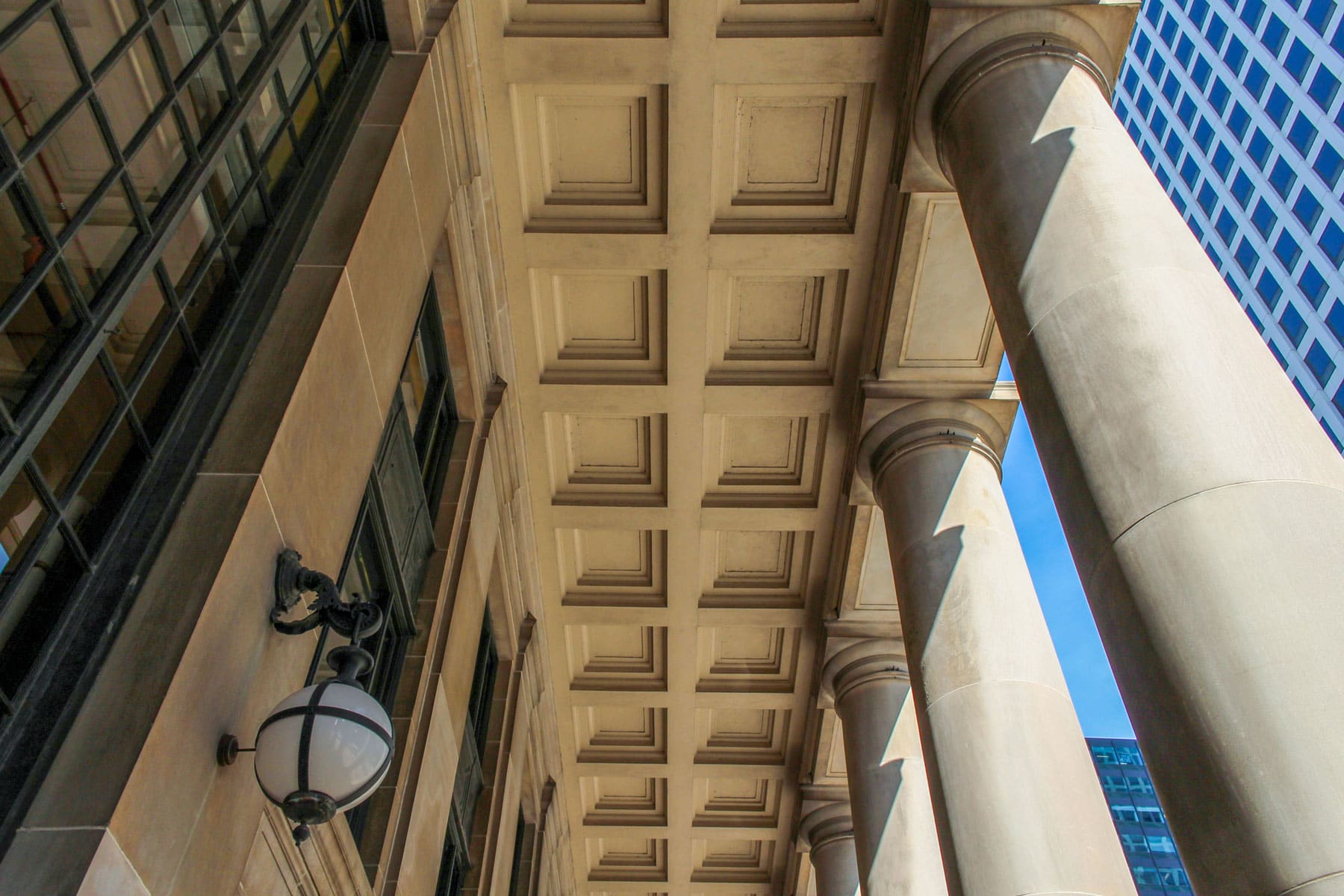 Architectural image capturing a row of stone columns and a coffered ceiling, with a vintage lamp on the wall. A blue skyscraper and clear sky in the background.