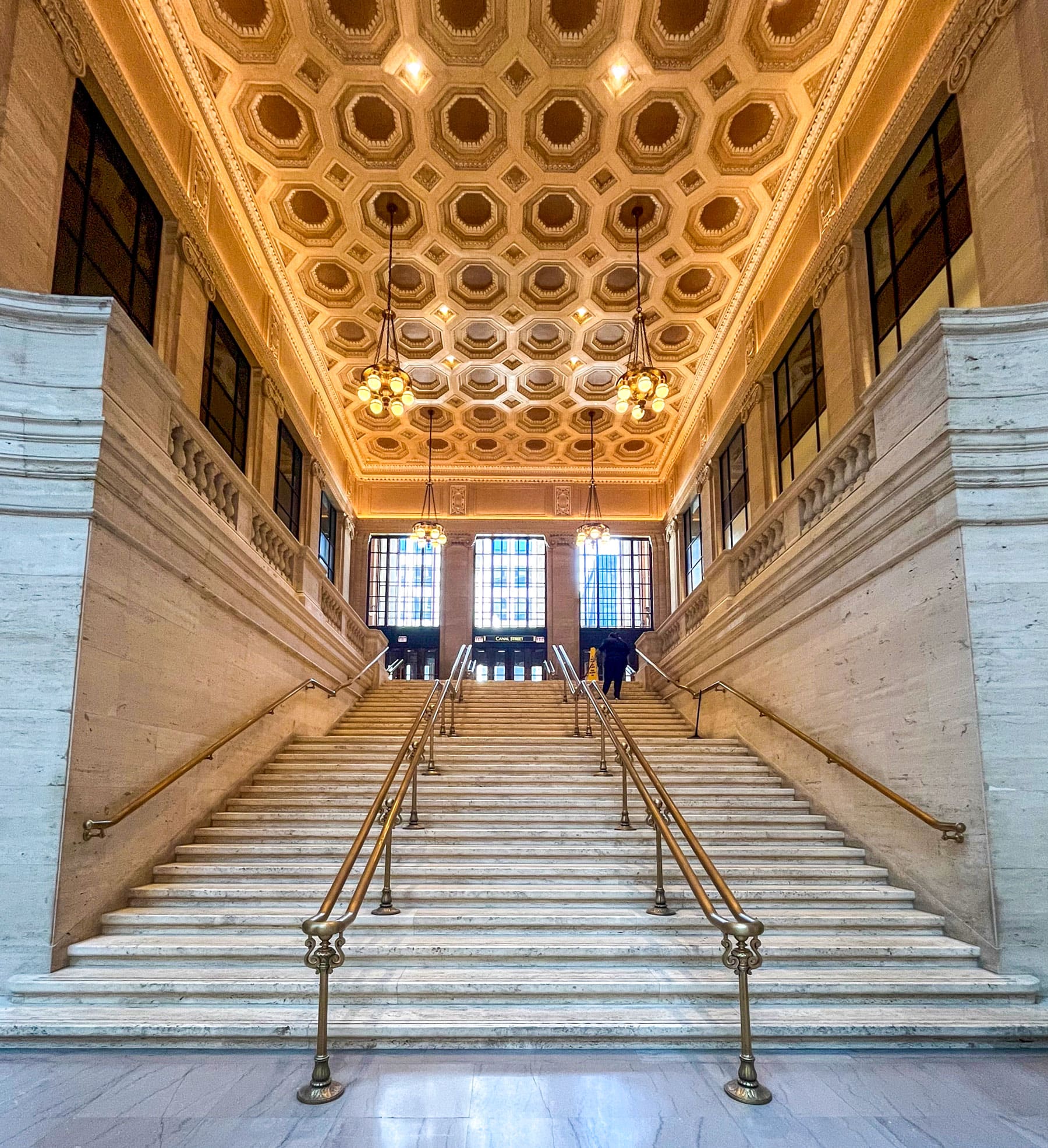 Grand staircase with ornate chandeliers and a hexagonal coffered ceiling. Tall windows illuminate the elegant, symmetrical stone architecture.