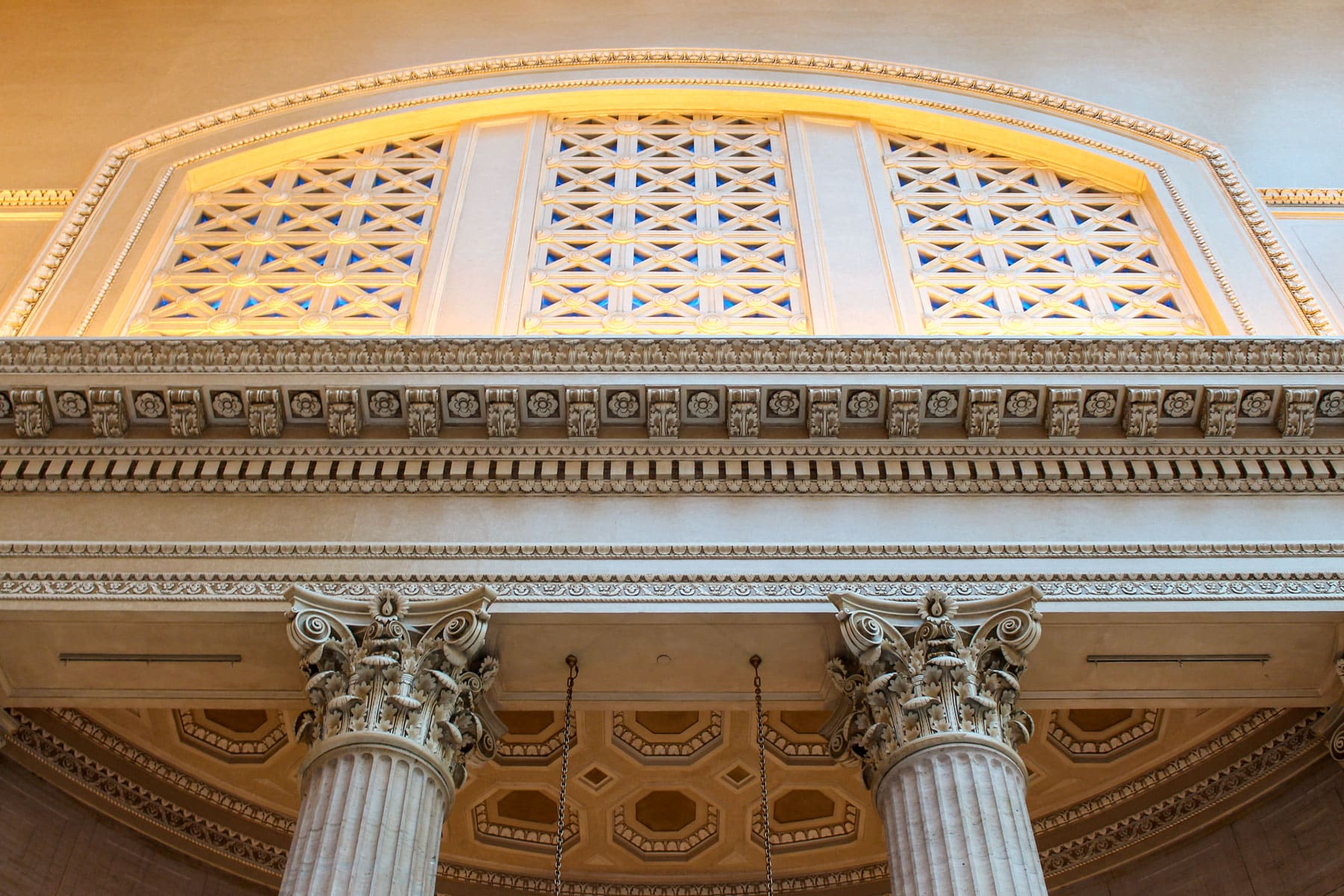 Ornate architectural detail featuring two Corinthian columns supporting an intricately carved entablature and decorative, sunlit arch window above.
