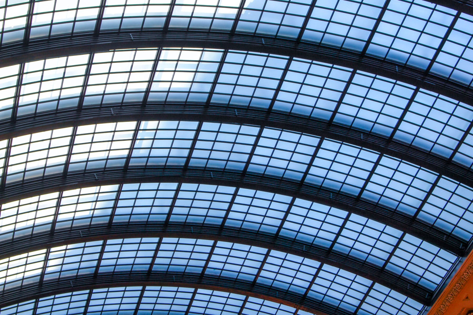 Curved glass roof with grid-like pattern, showcasing blue sky. The structure conveys elegance and modern architectural design.