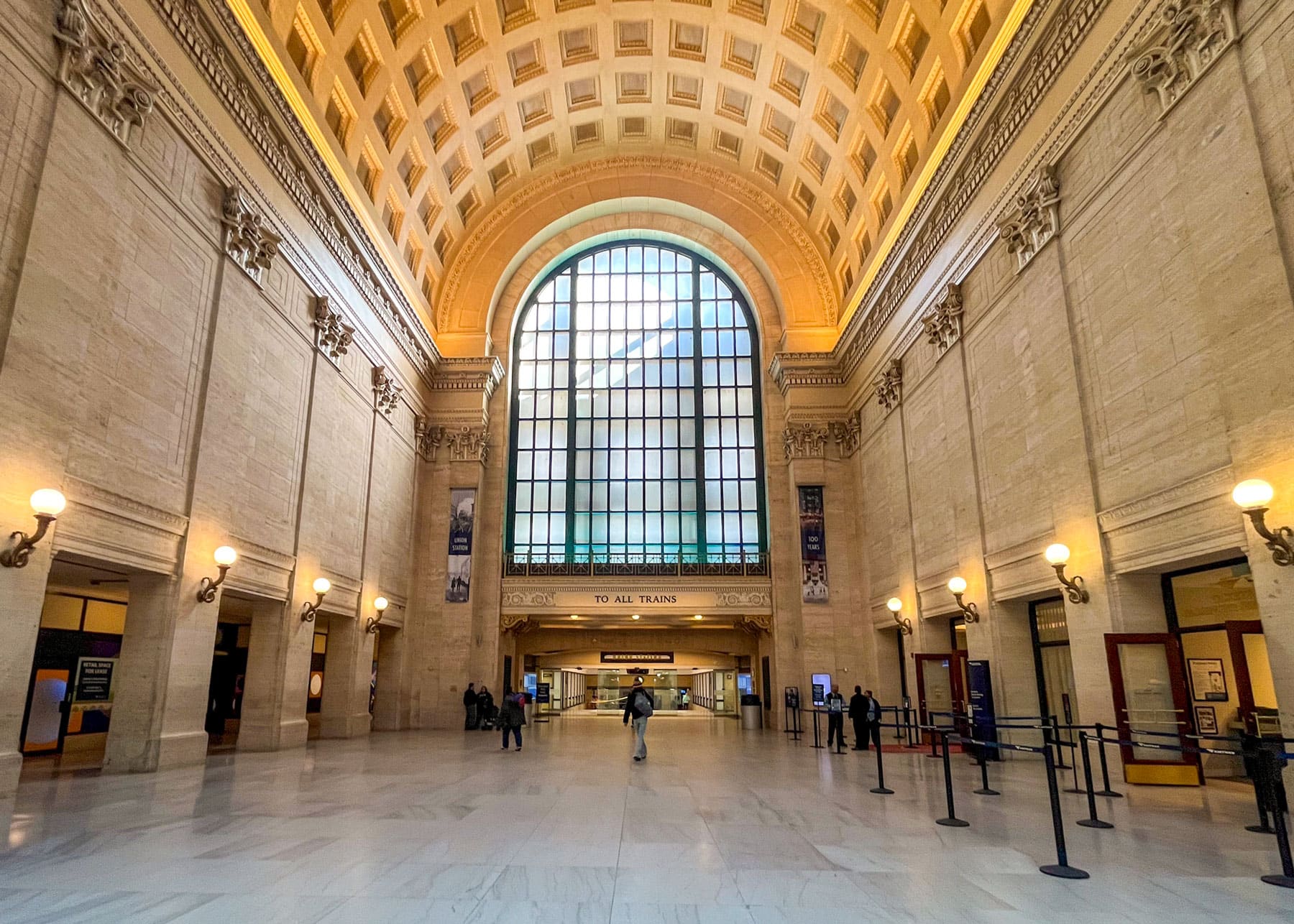 Grand, high-ceilinged train station hall with large arched window. Warm lighting, ornate detailing. Few people walking below sign to all trains.