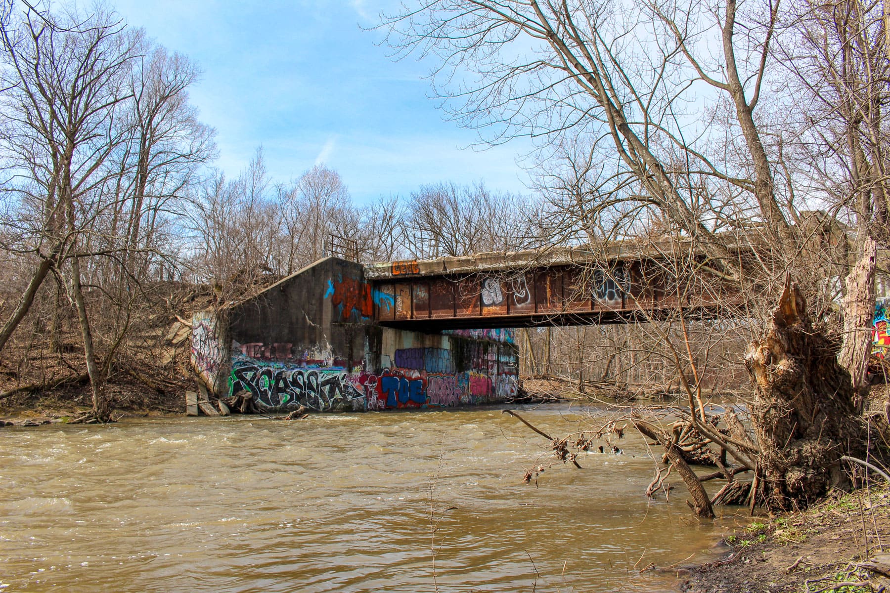 Abandoned graffiti-covered bridge over a muddy river, surrounded by leafless trees. The scene conveys a sense of decay and urban art.