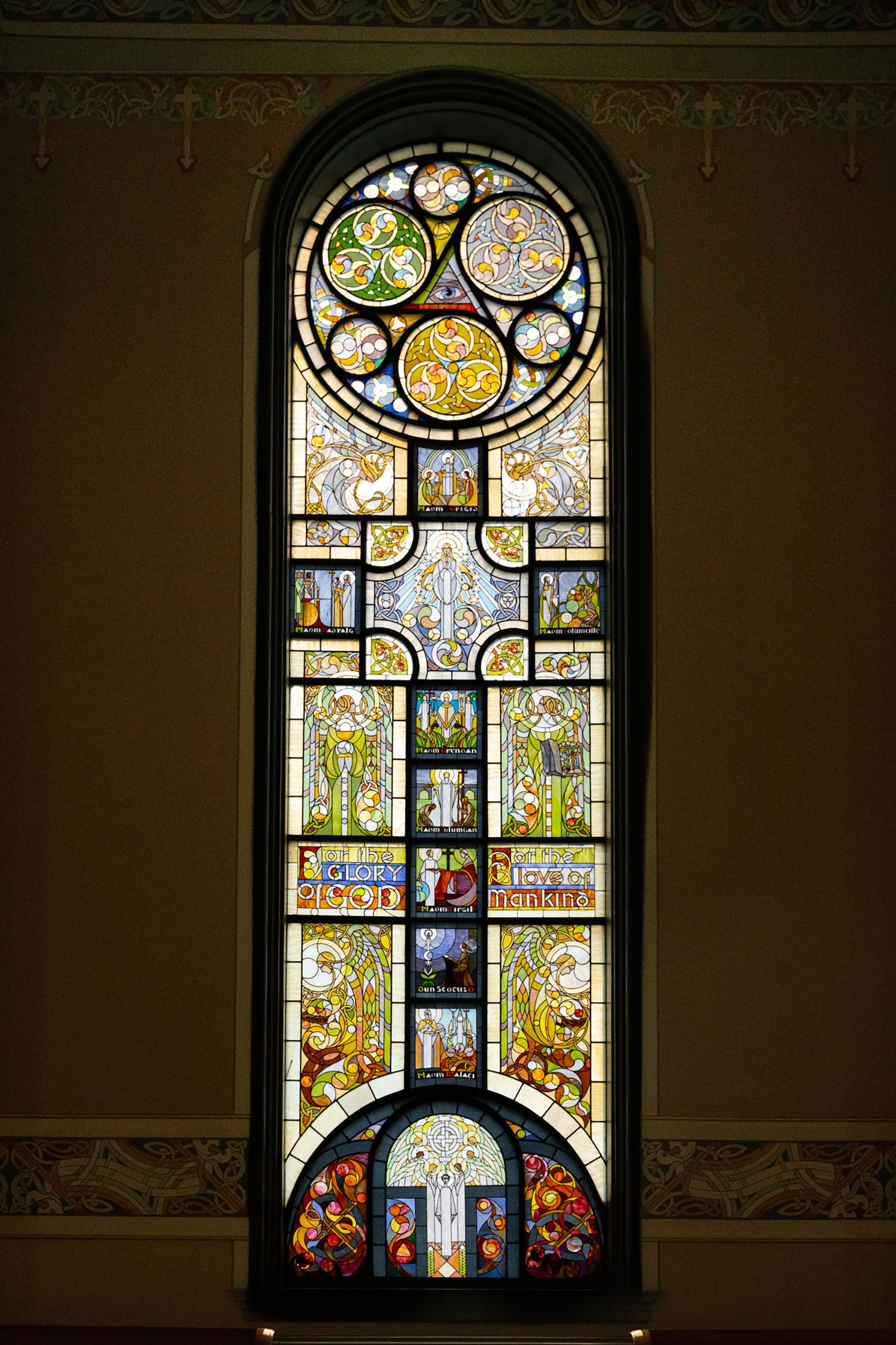 An arched Celtic Revival stained glass window featuring intricate interlocking circular patterns, colorful knotwork, and multi-paneled religious imagery.